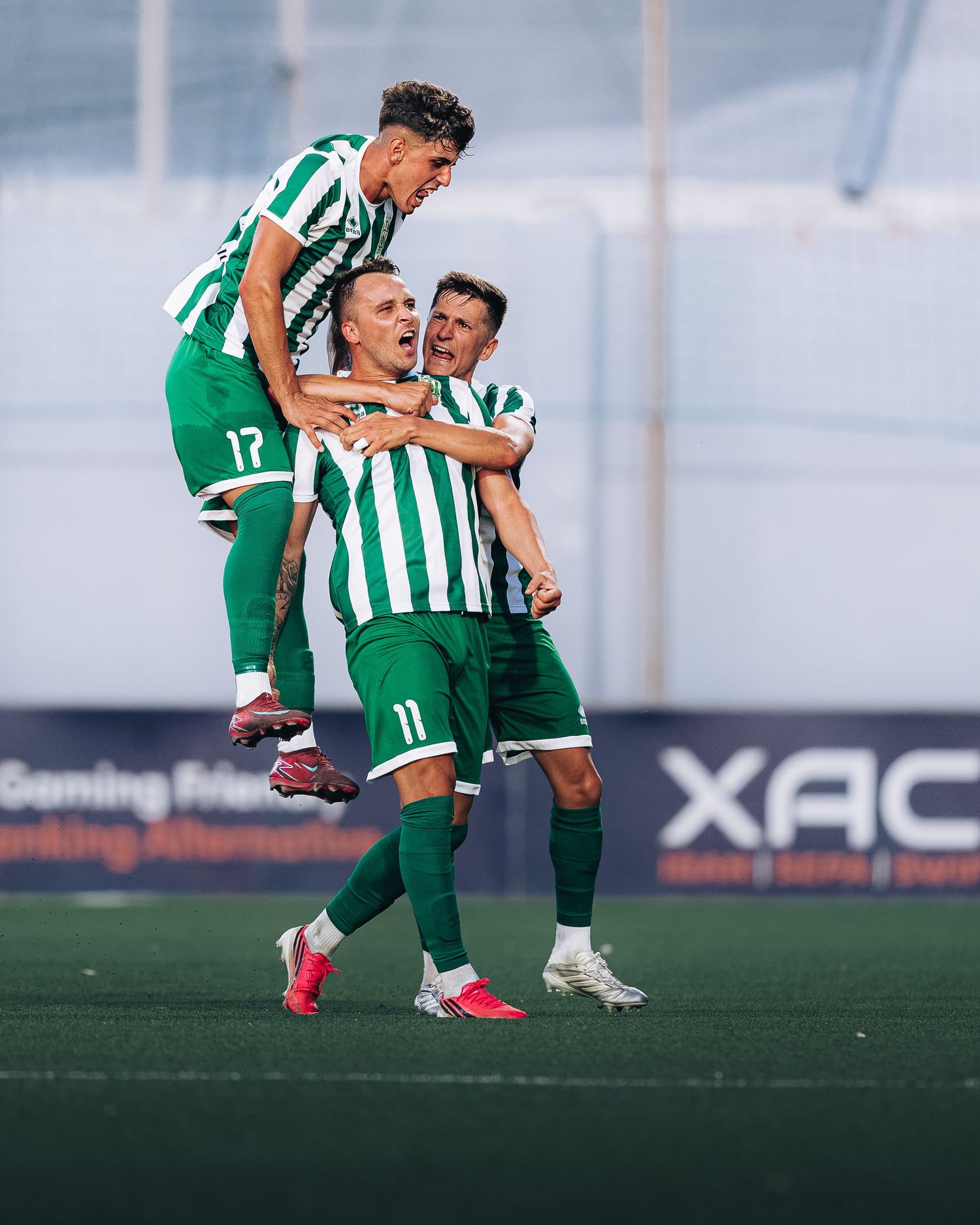 Three football players in green and white jerseys celebrating a goal together on the pitch.