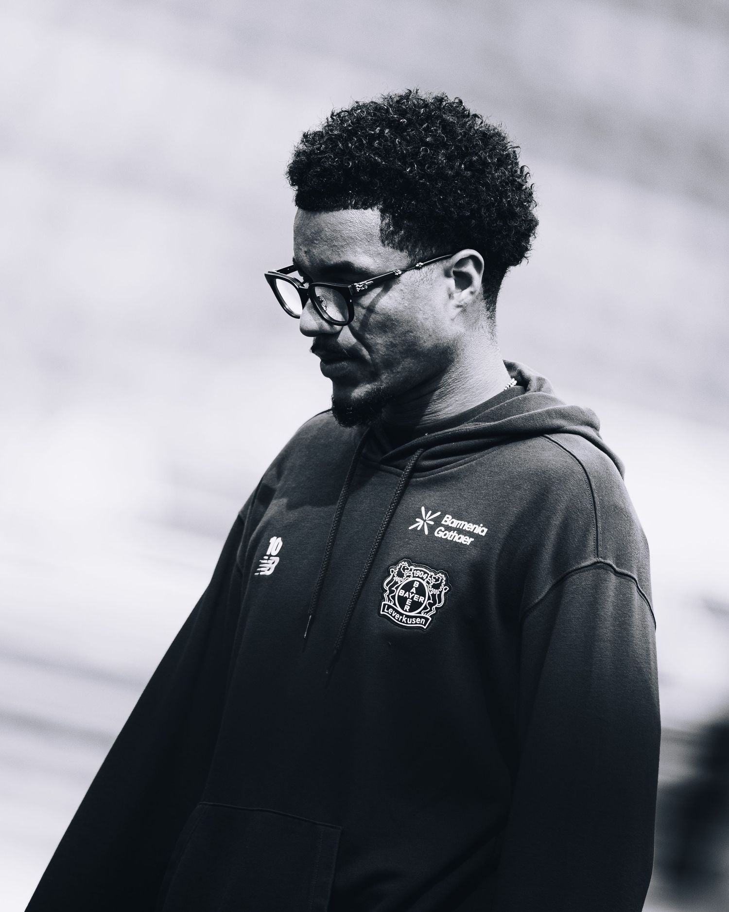 Black and white portrait of a man wearing glasses and a dark Bayer Leverkusen hoodie looking downward.