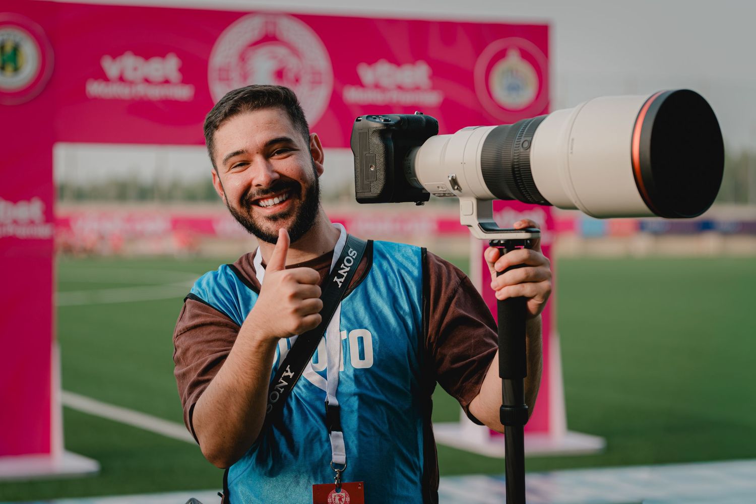Smiling sports photographer gives thumbs up holding large telephoto lens at soccer stadium.