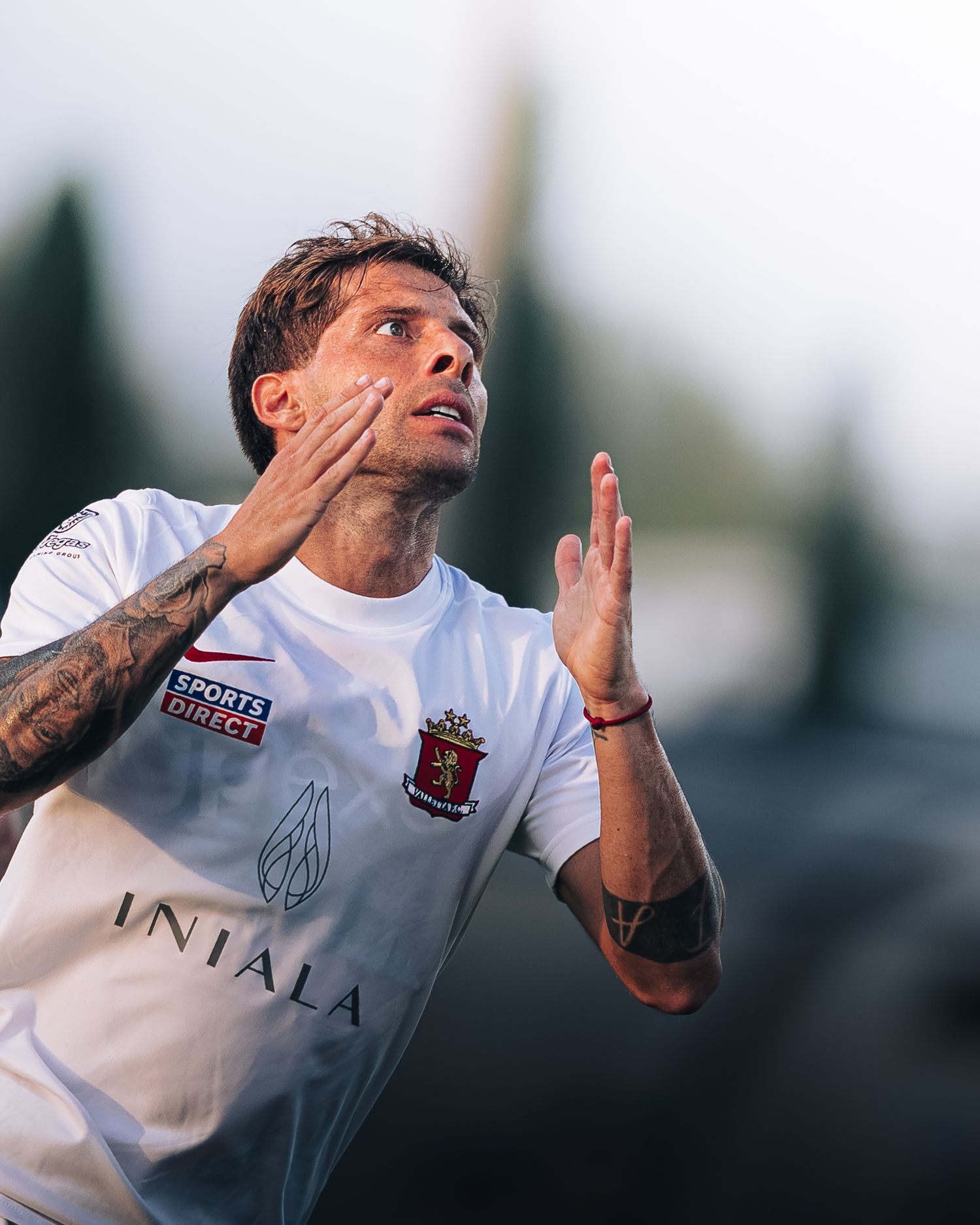 Tattooed footballer in white Injala sponsored shirt looking skyward with hands raised.