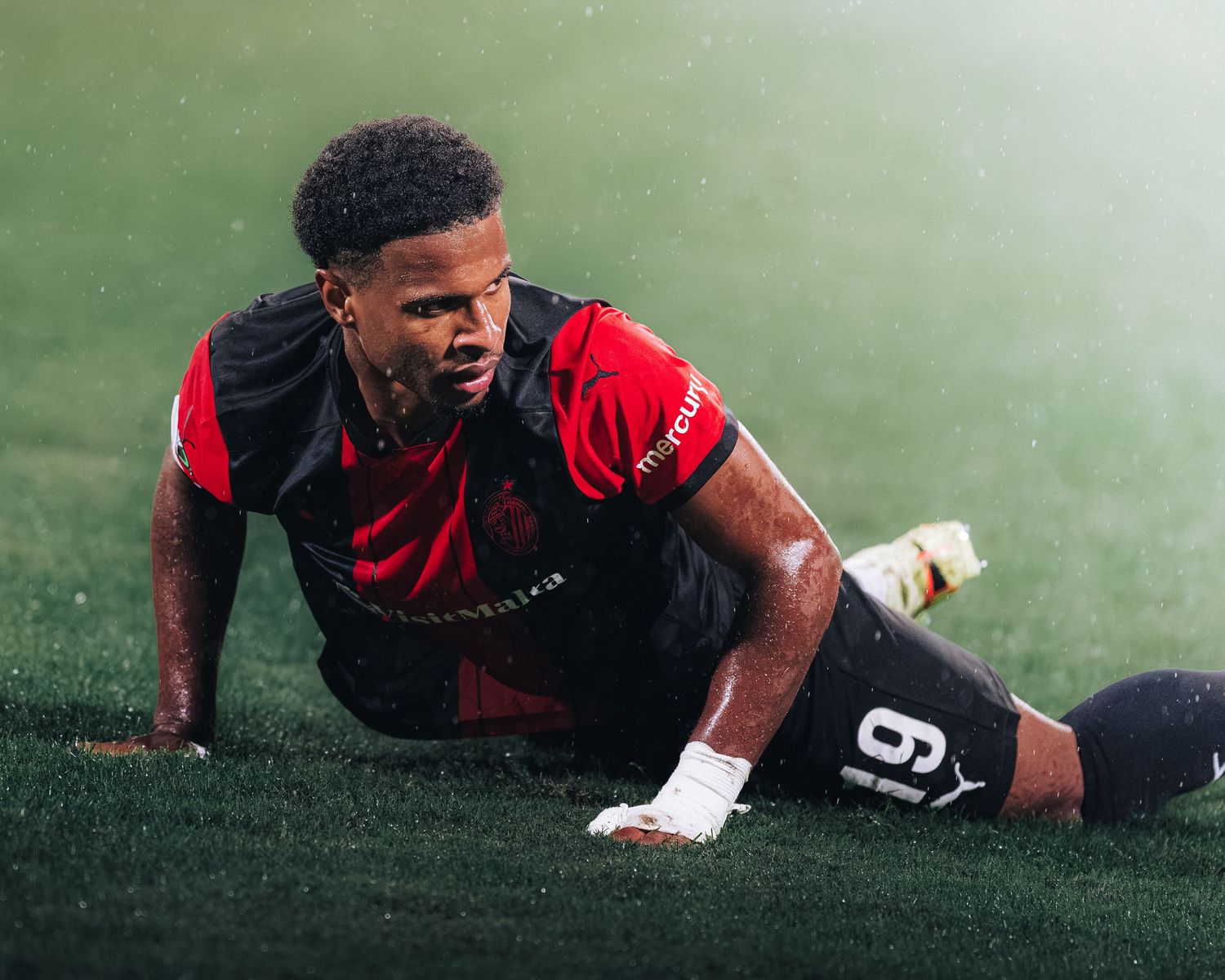 Soccer player in red and black jersey number 9 slides on wet grass during match.
