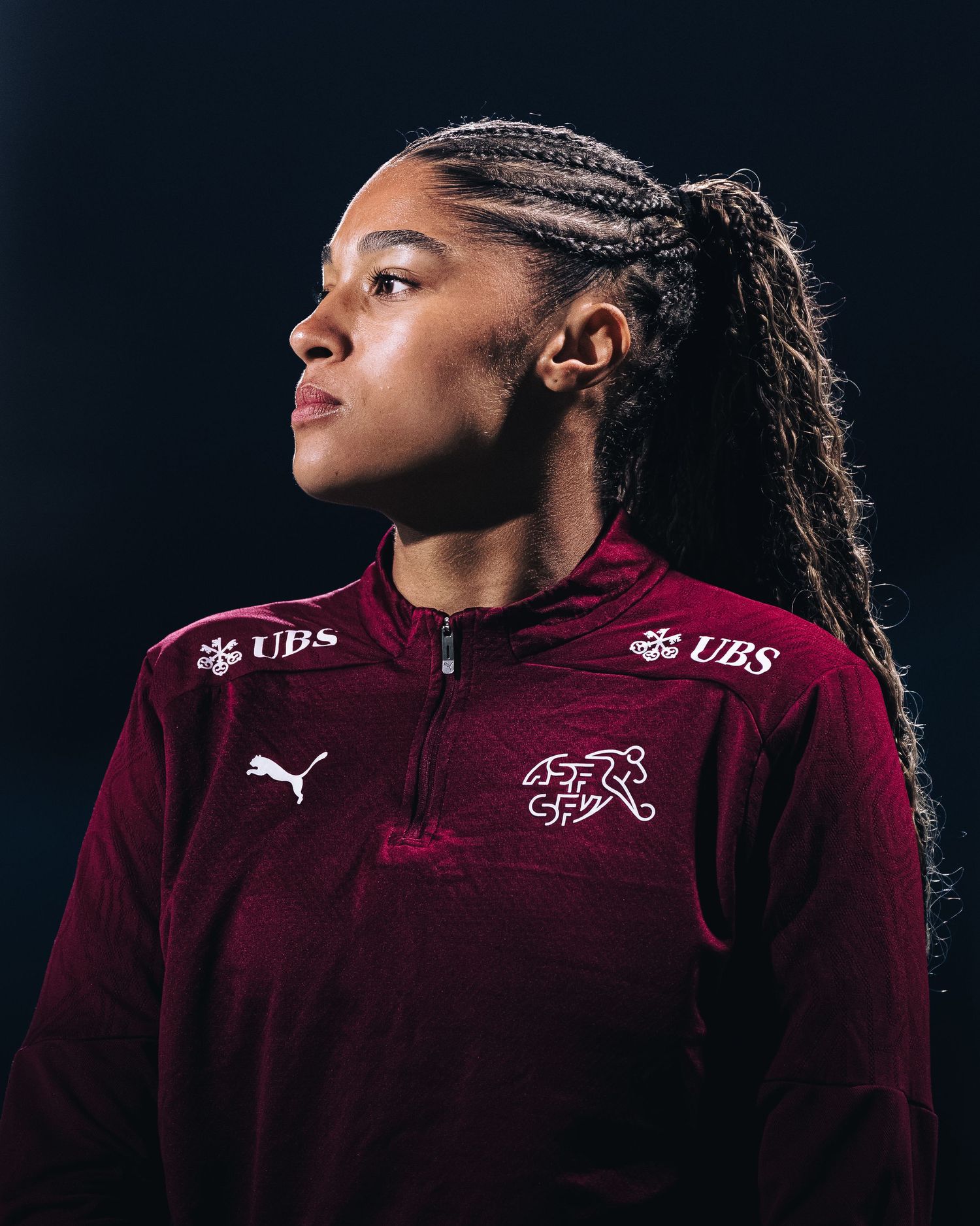 Female soccer player in maroon Swiss national team PUMA jersey with UBS sponsor logo poses against dark background.