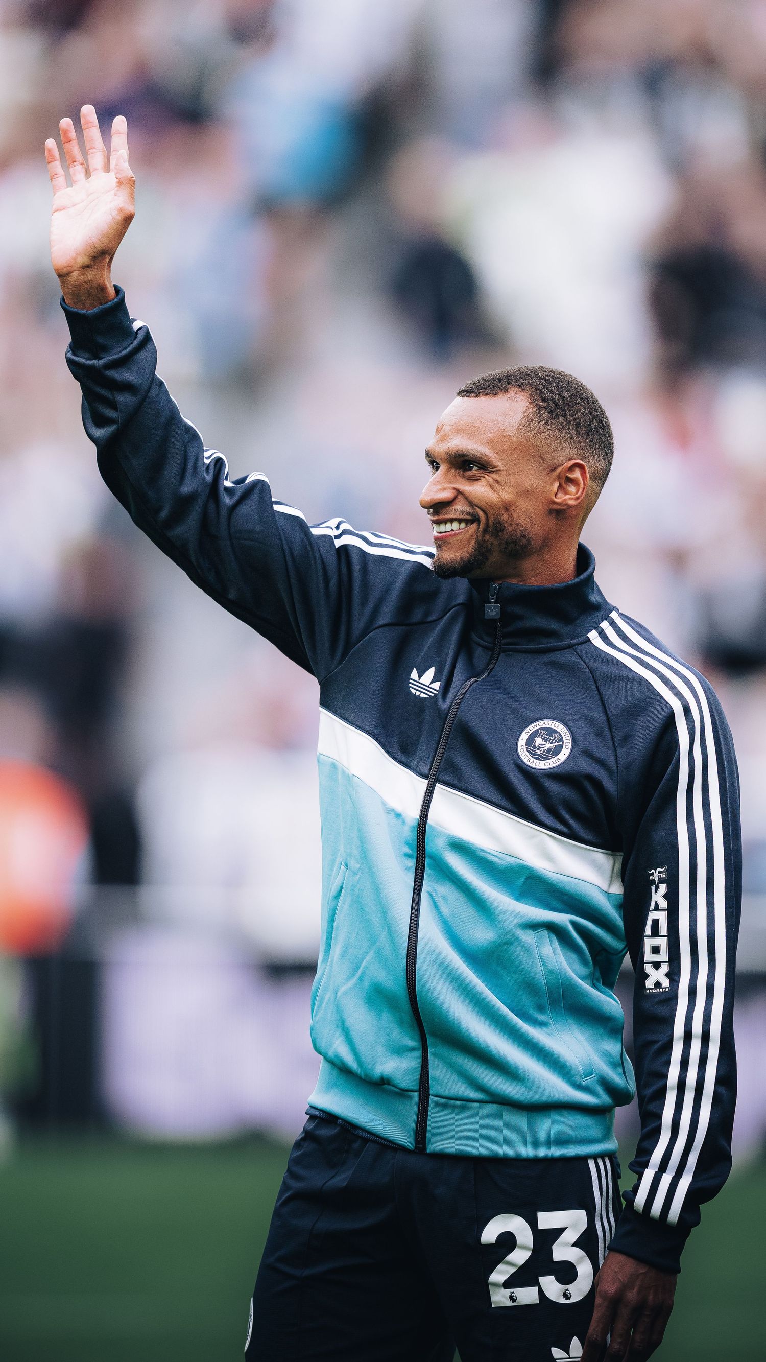Smiling footballer in Adidas tracksuit number 23 waves to fans at stadium.