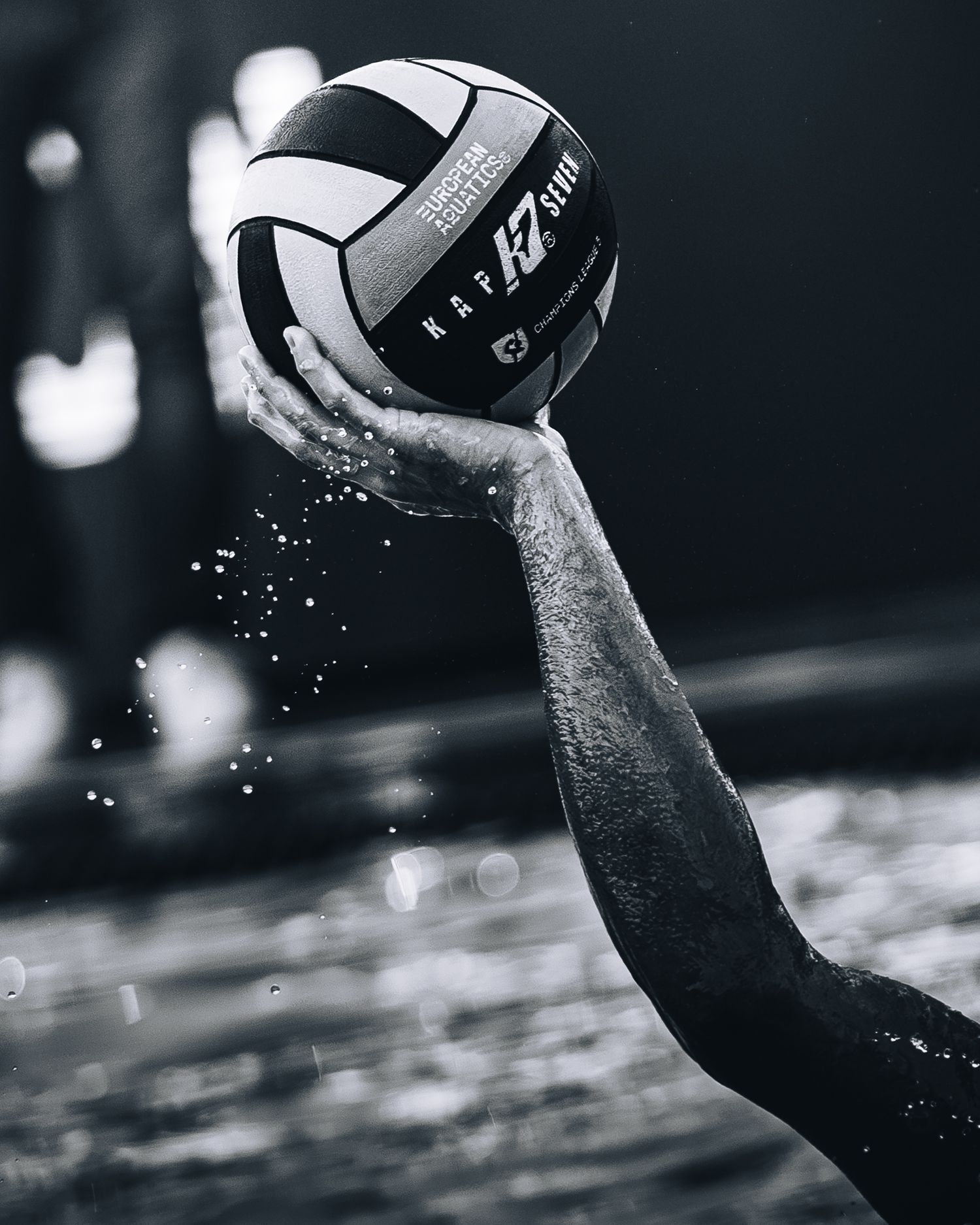 Black and white close-up of hand gripping KAP7 water polo ball rising from splashing water.