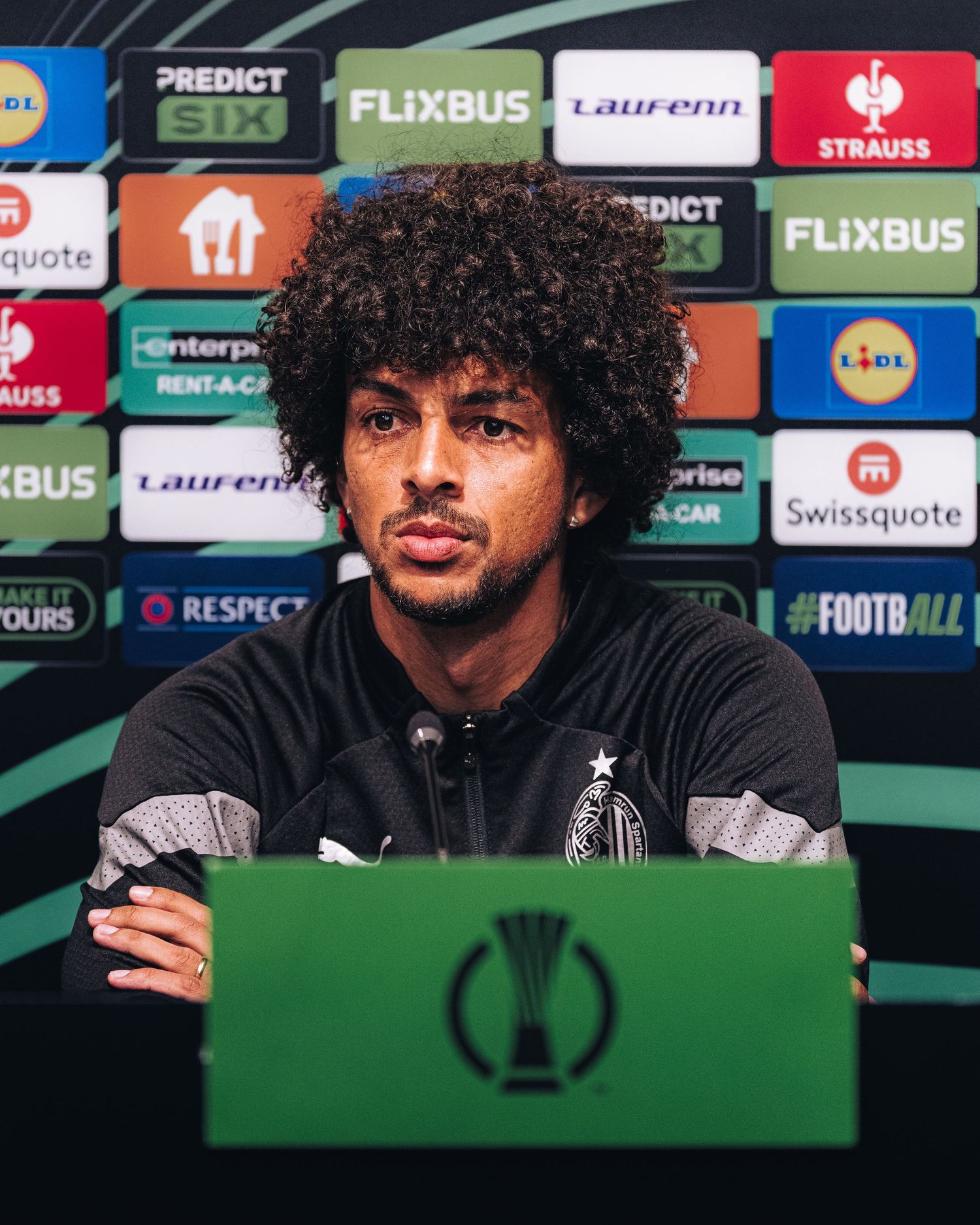 Curly-haired footballer in black kit speaking at UEFA Conference League press conference podium.