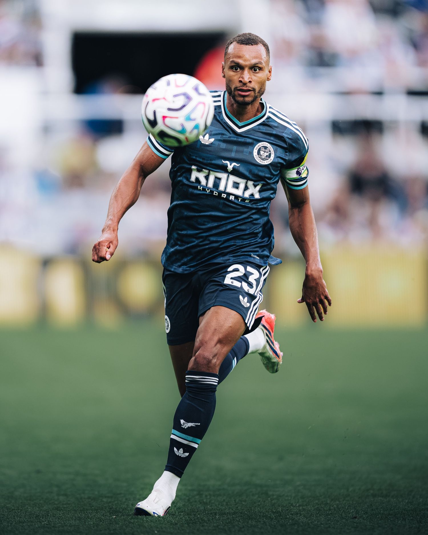Footballer in dark Knox Hydrate sponsored kit controls ball during sunny match.