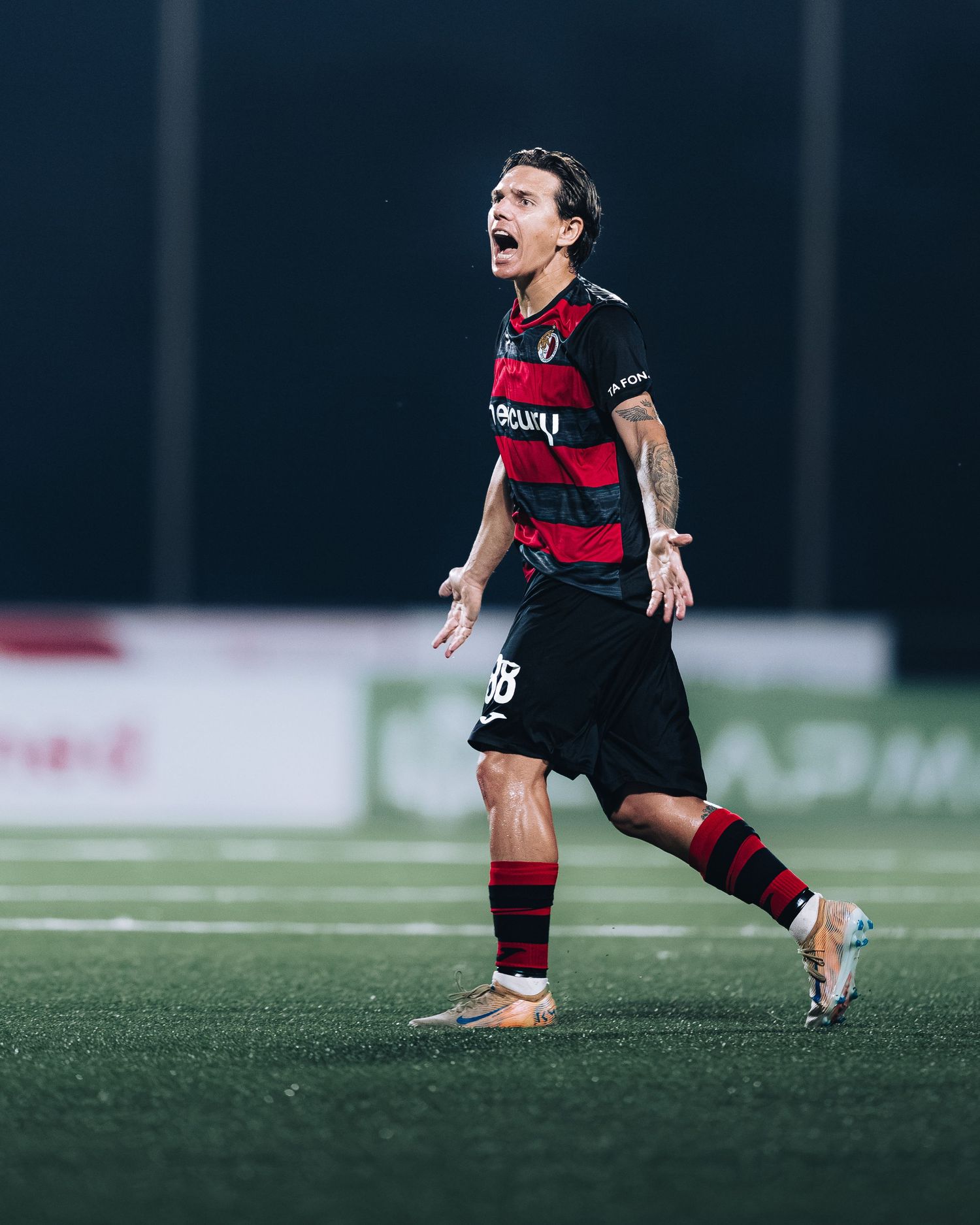 Soccer player in red and black striped uniform reacts emotionally on illuminated night field.