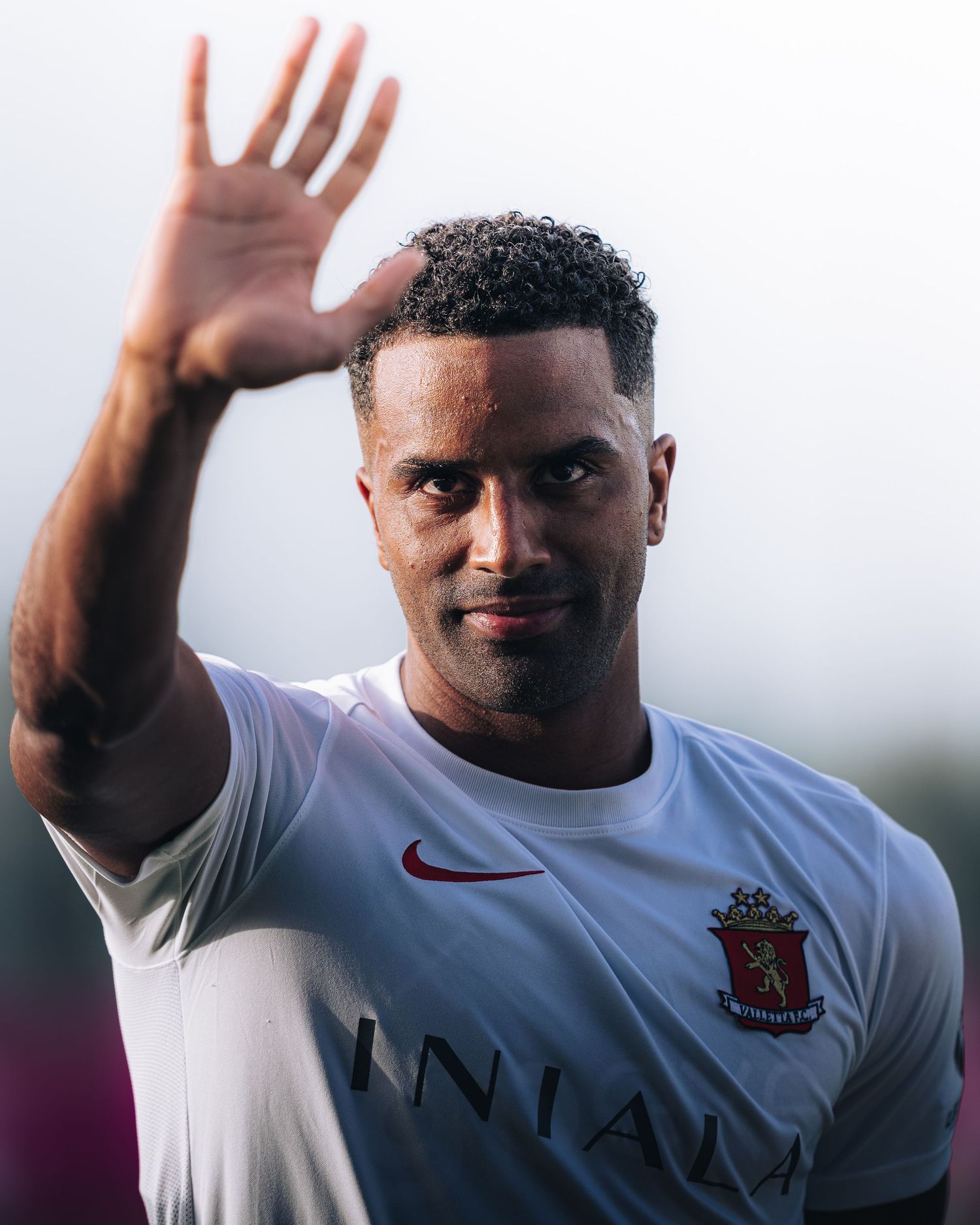 Soccer player in white Valletta FC Nike jersey smiling and waving to fans.