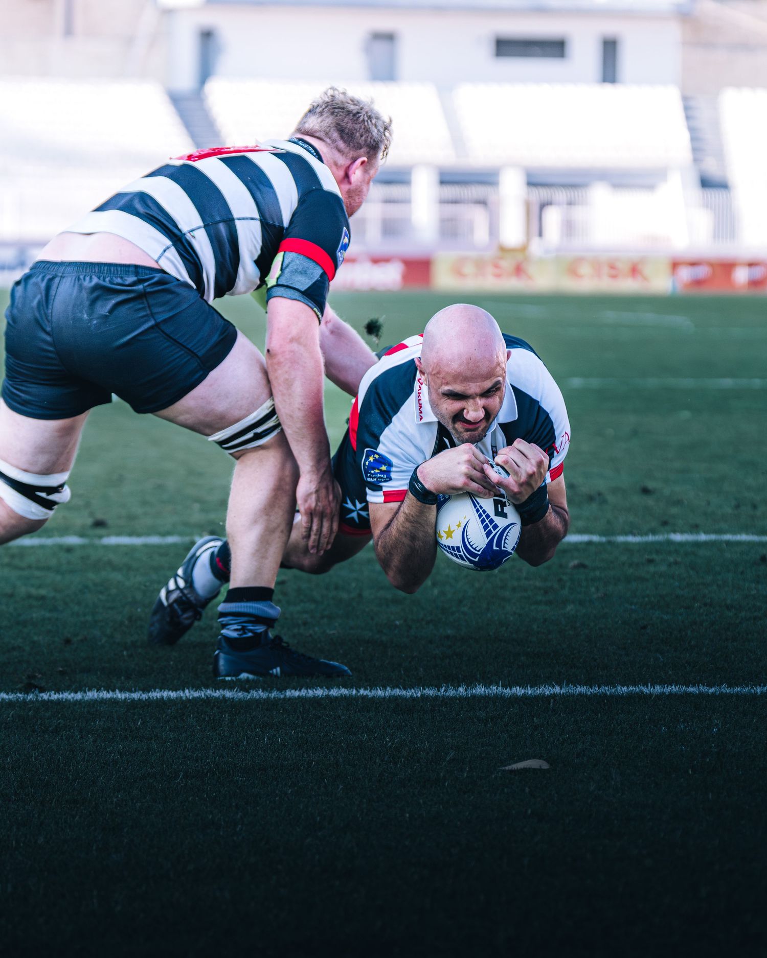 Bald rugby player diving to score try while opponent attempts tackle in stadium.
