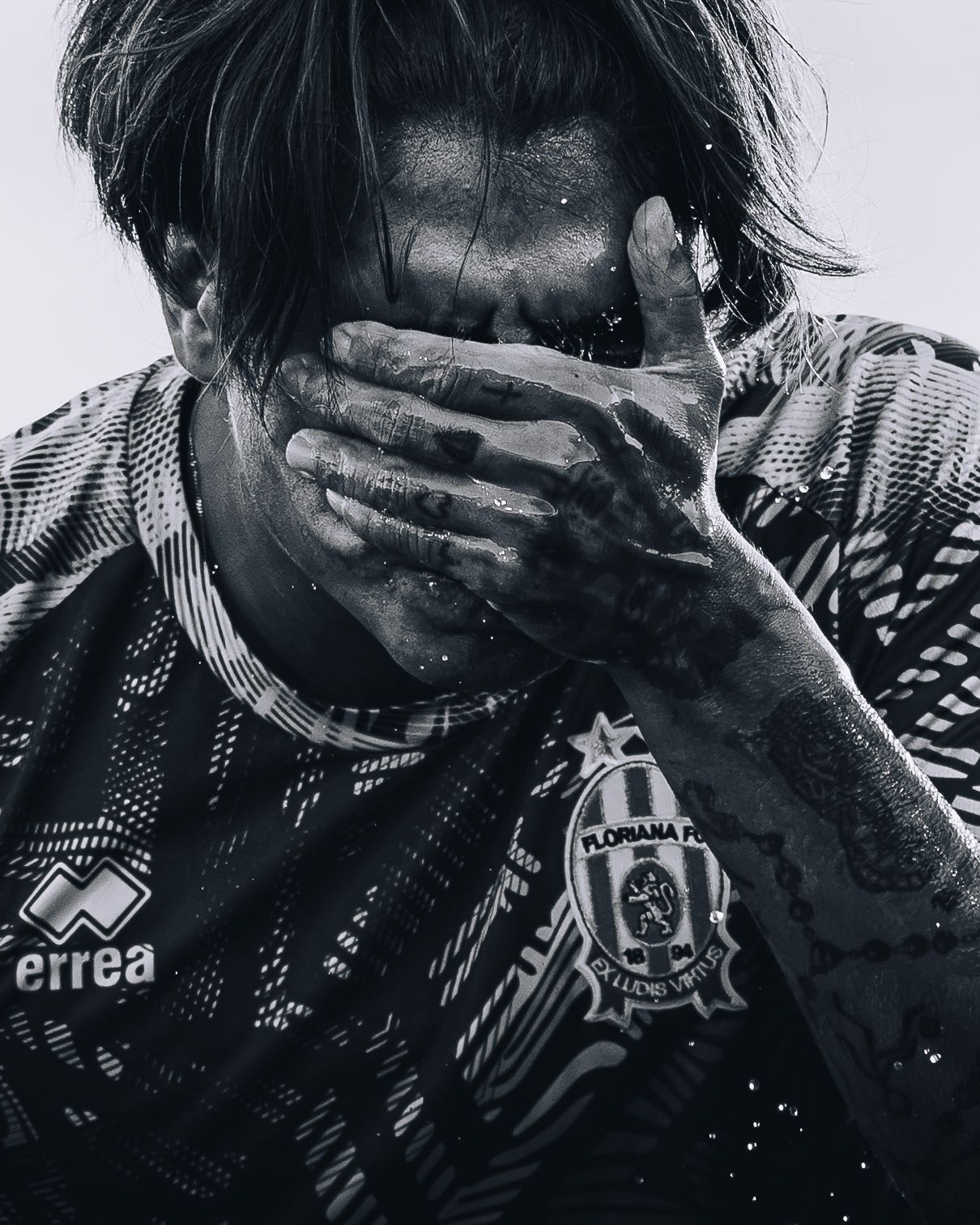 Black and white close-up of muddy soccer player covering face with hand wearing Errea kit.