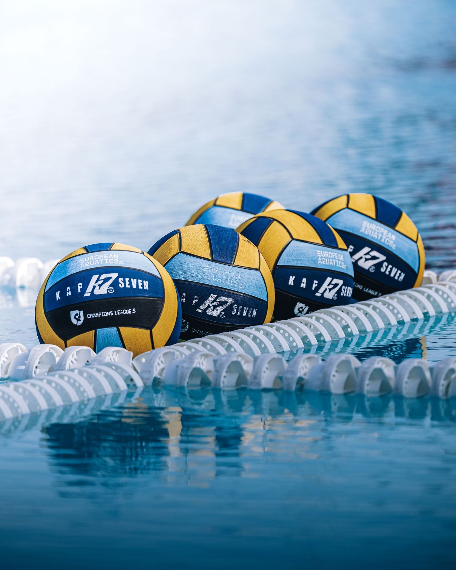 Four KAP7 water polo balls floating on pool lane line in blue water during competition.