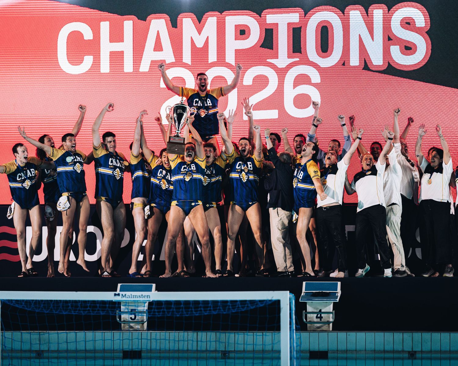 Water polo team celebrating championship victory under Champions 2026 banner poolside.