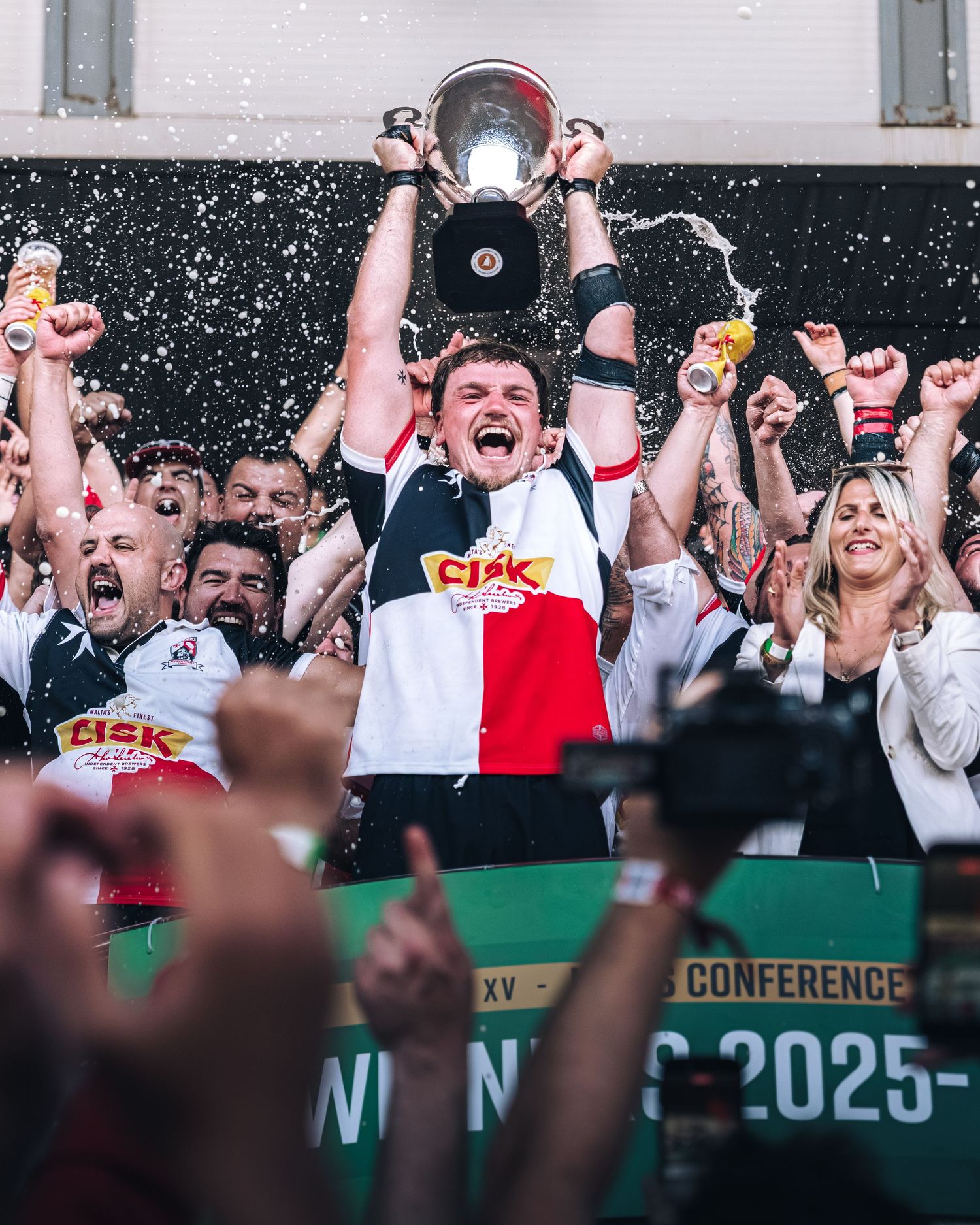 Rugby team captain lifts trophy in celebration surrounded by cheering teammates and confetti.