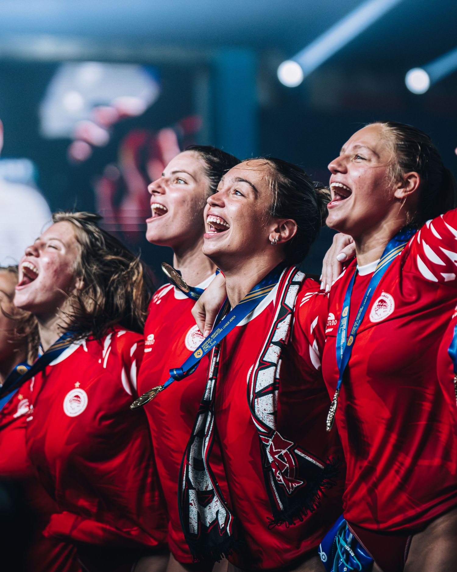 Female water polo team in red jerseys joyfully celebrating championship win with medals.