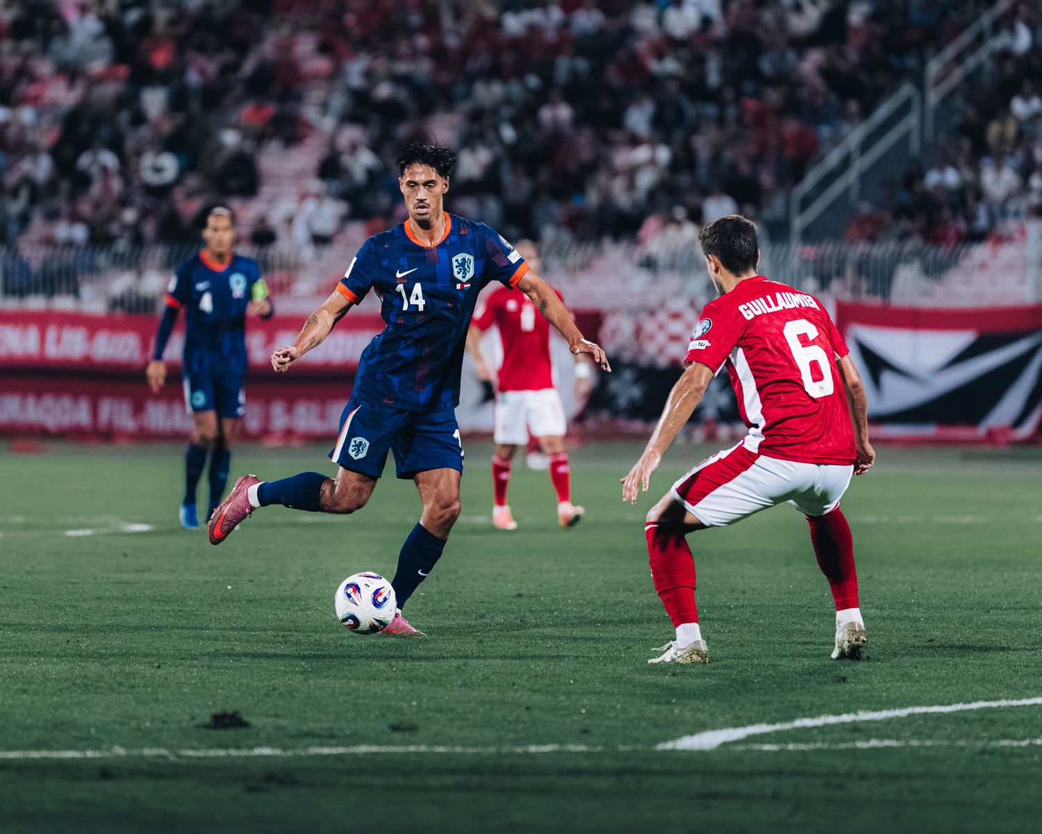 Football player number 14 in blue kit dribbling past defender number 6 in red and white during a match.