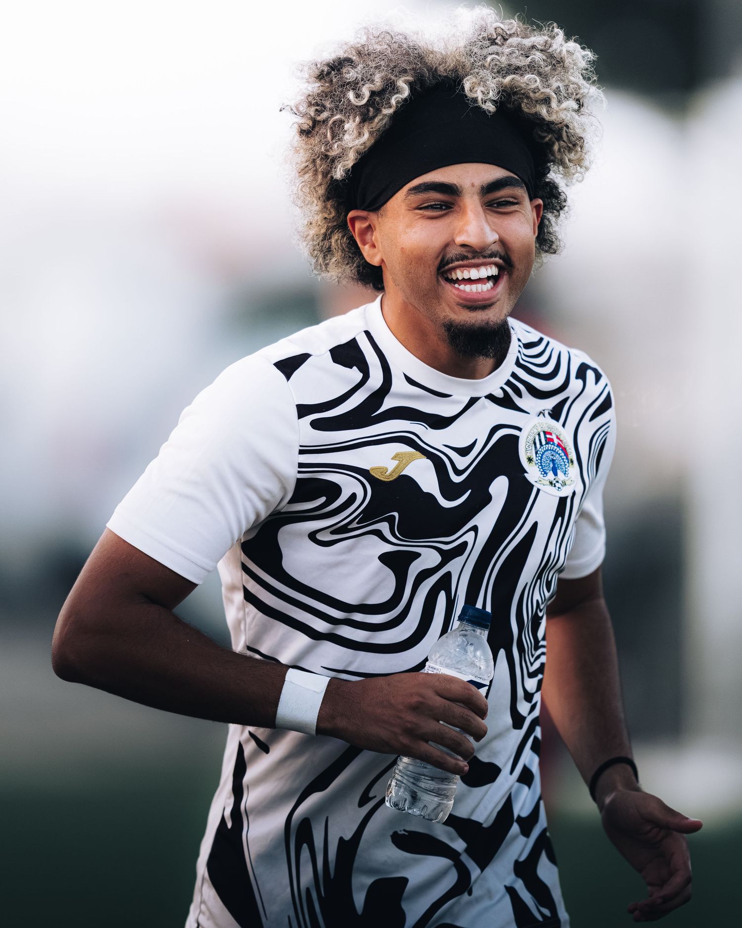 Smiling male athlete with curly hair wearing black and white abstract print jersey holds water bottle outdoors.