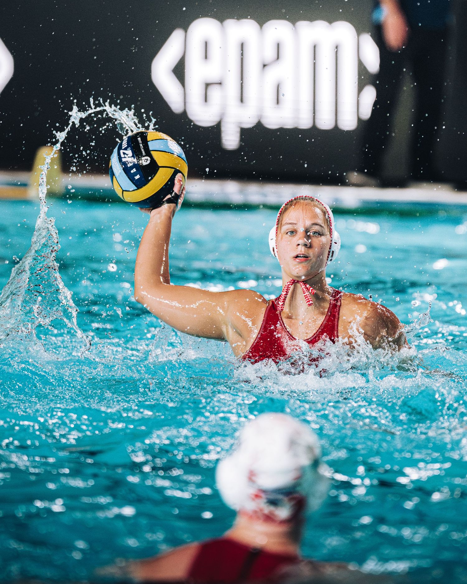 Female water polo player in red cap about to throw the ball during a match with EPAM sponsorship.