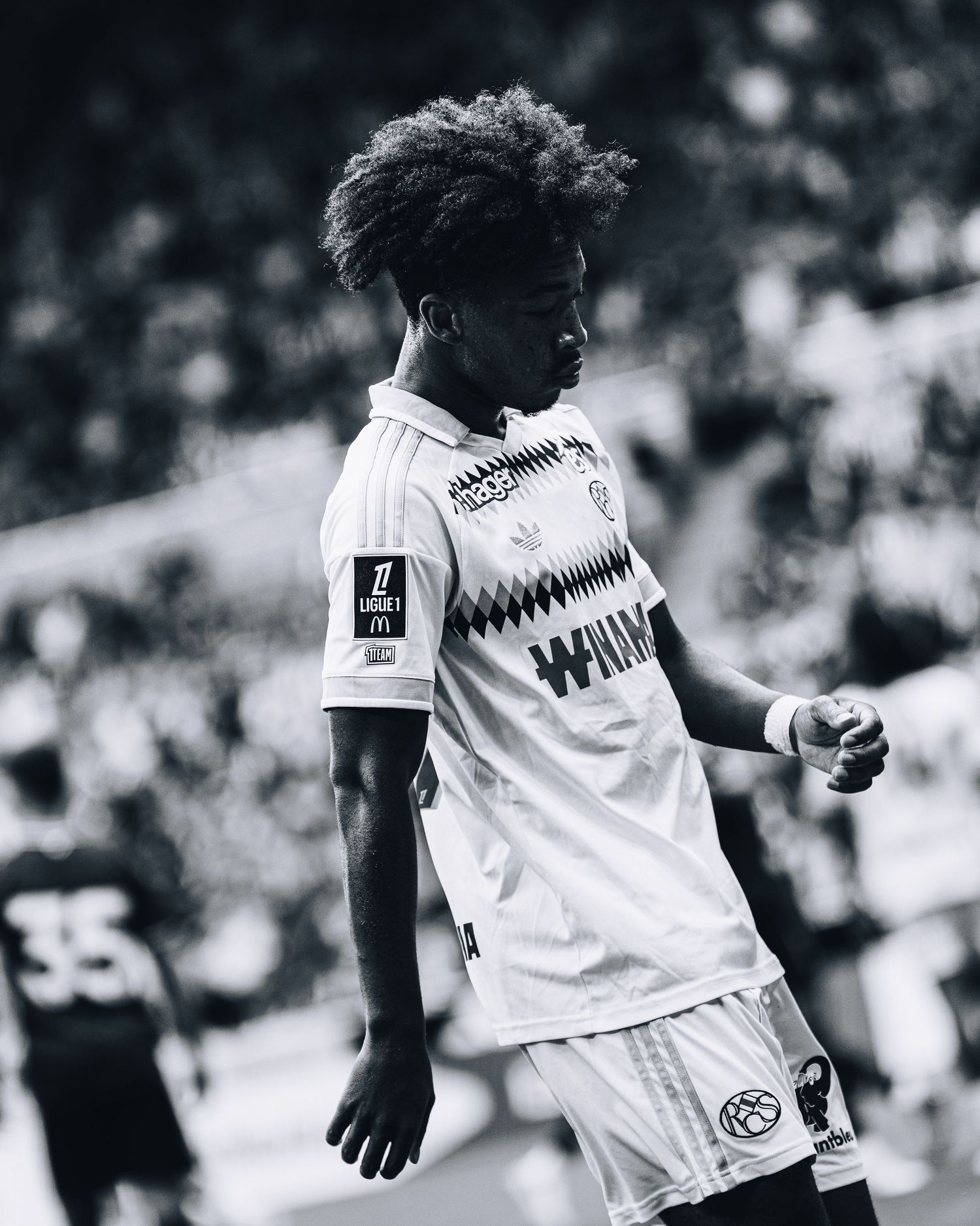 Black and white image of young curly-haired soccer player in Winmau sponsored white jersey on field.