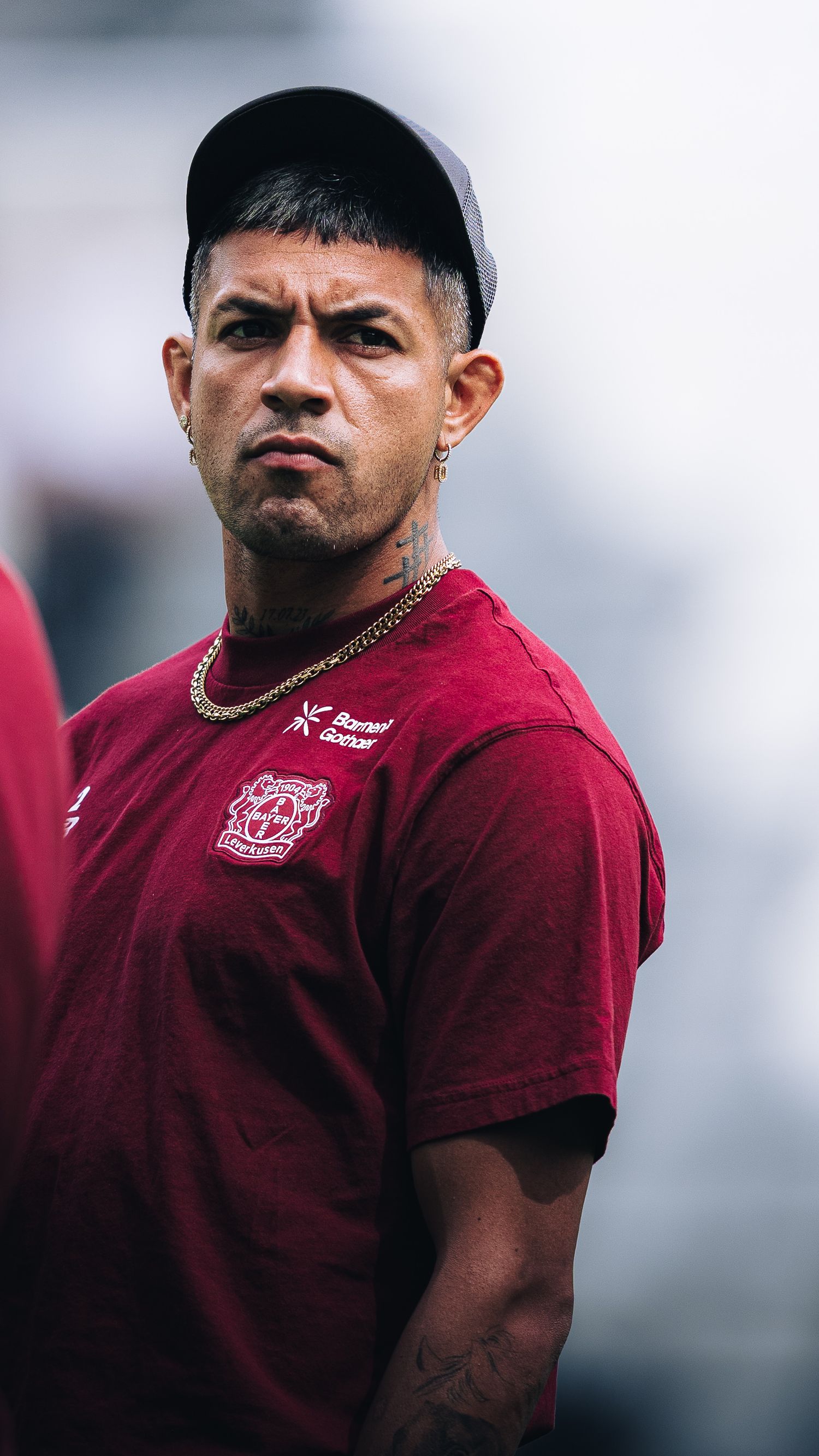 Tattooed male soccer player in maroon team shirt and cap with gold chain looks sideways.