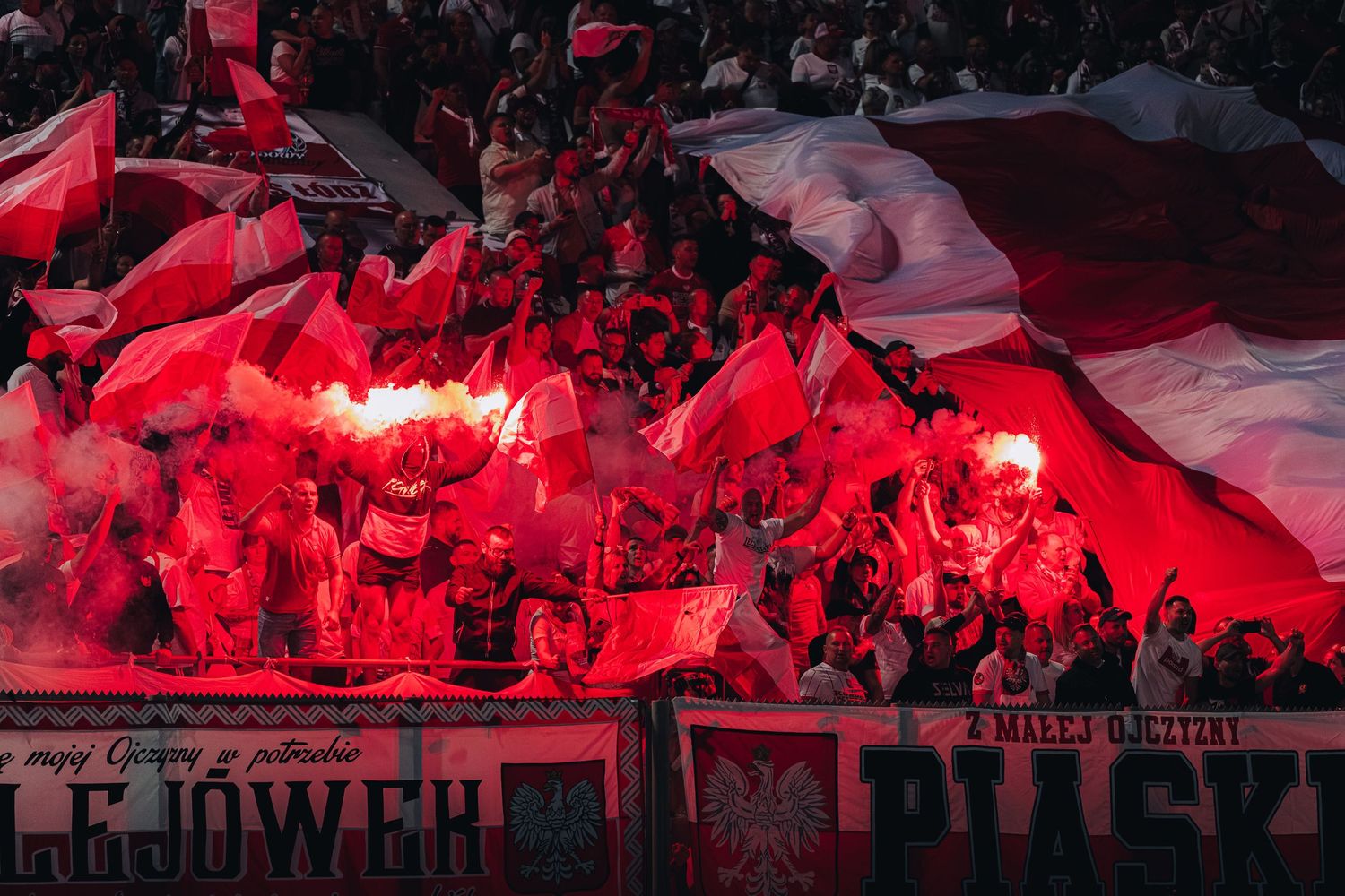 Polish football ultras with red smoke flares and large red and white national flag in stands.