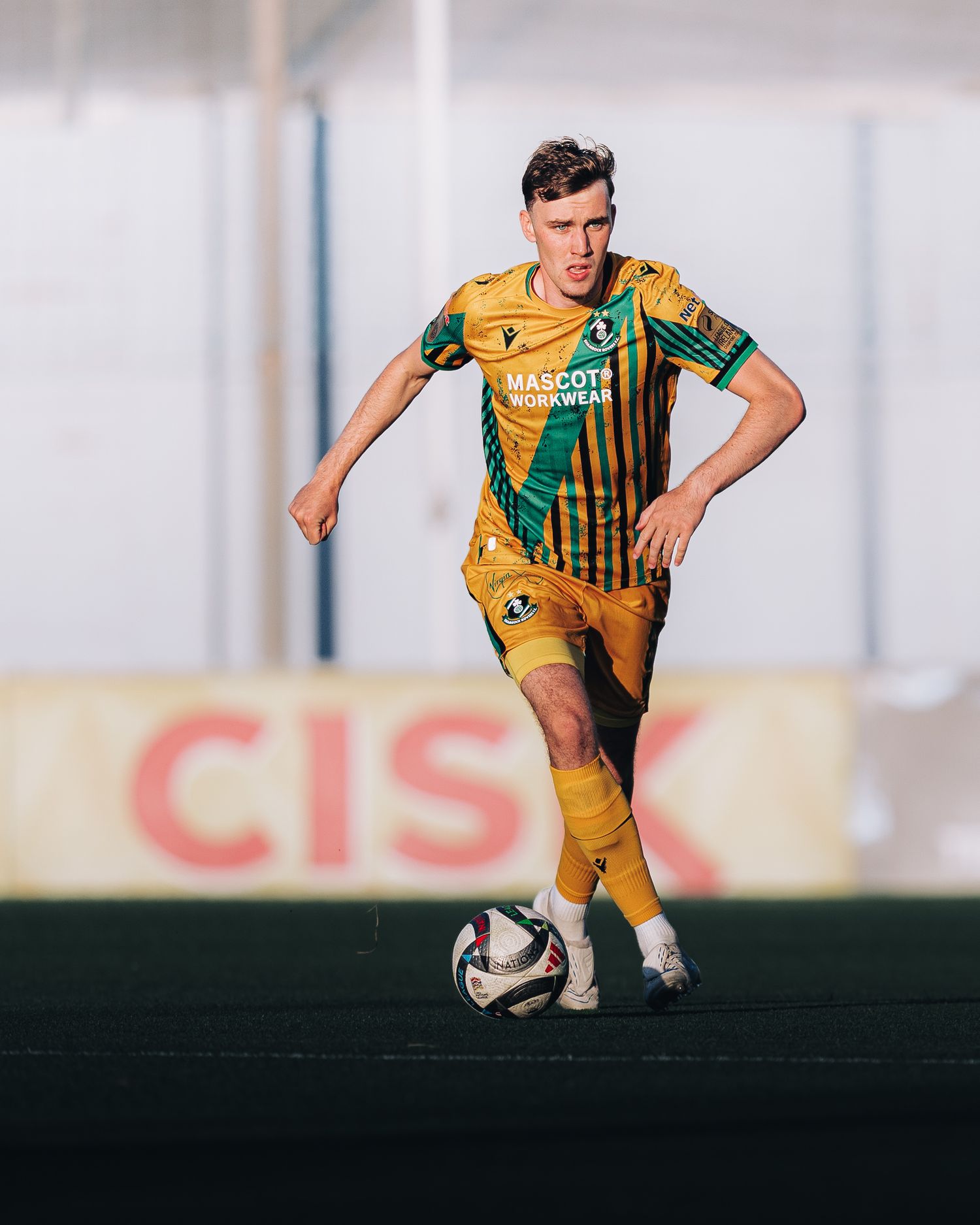 Soccer player in yellow and green kit dribbling ball on field with CISK advertising board in background.