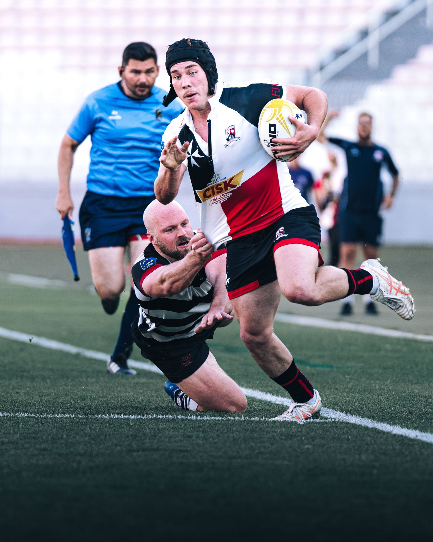 Rugby player in white kit breaks through a tackle during an intense match on a green field.