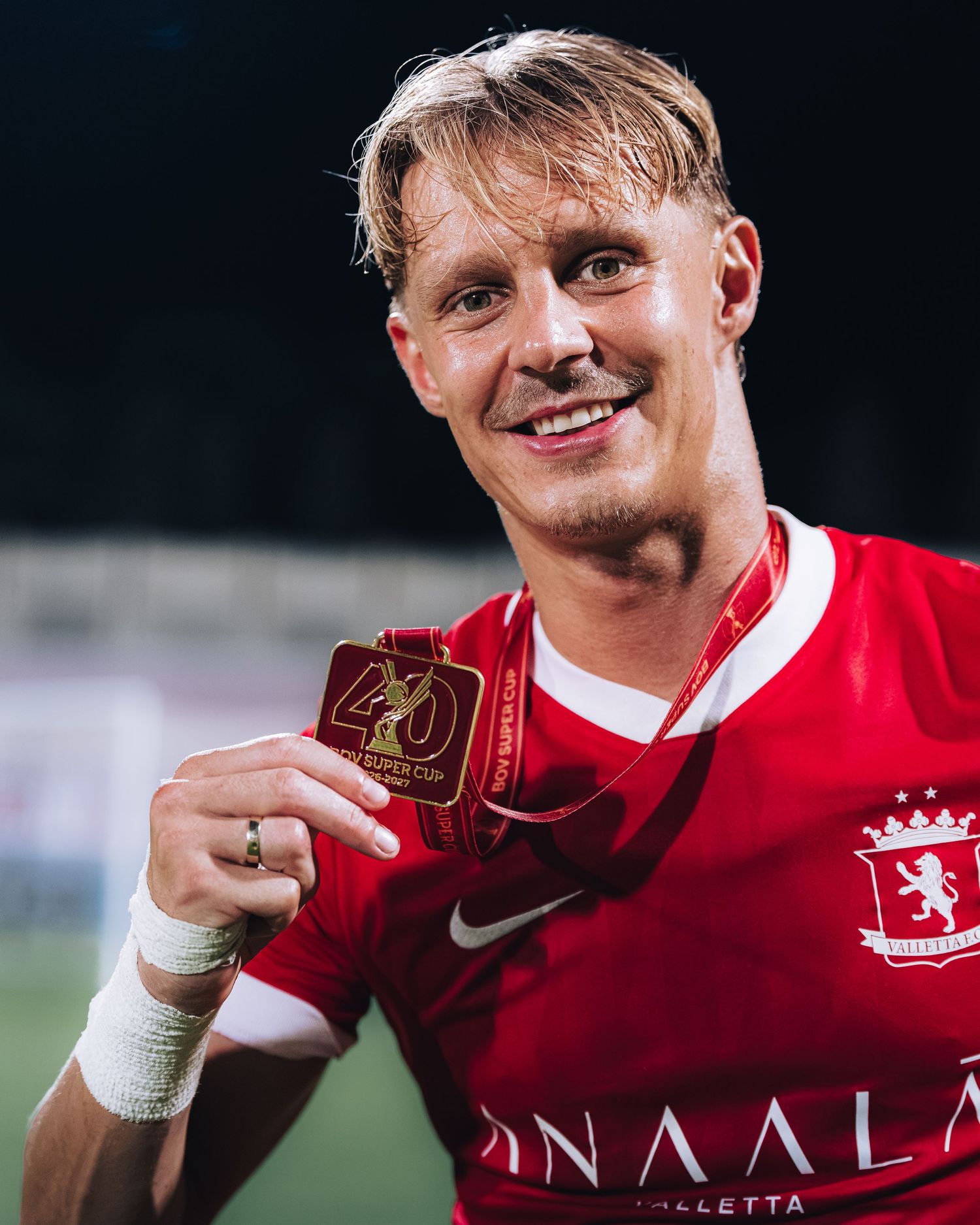 Smiling blond soccer player in red Valletta FC jersey proudly holding gold medal.