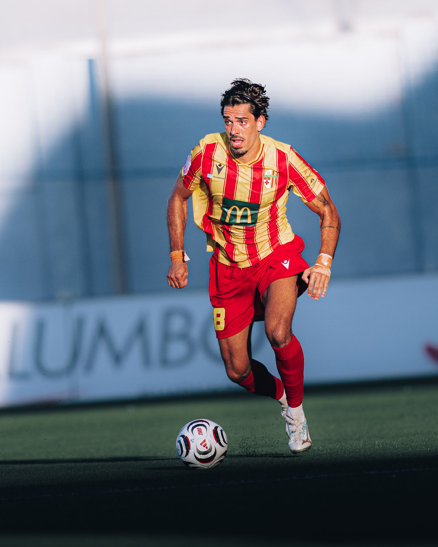 Soccer player in red and yellow striped kit dribbles ball.
