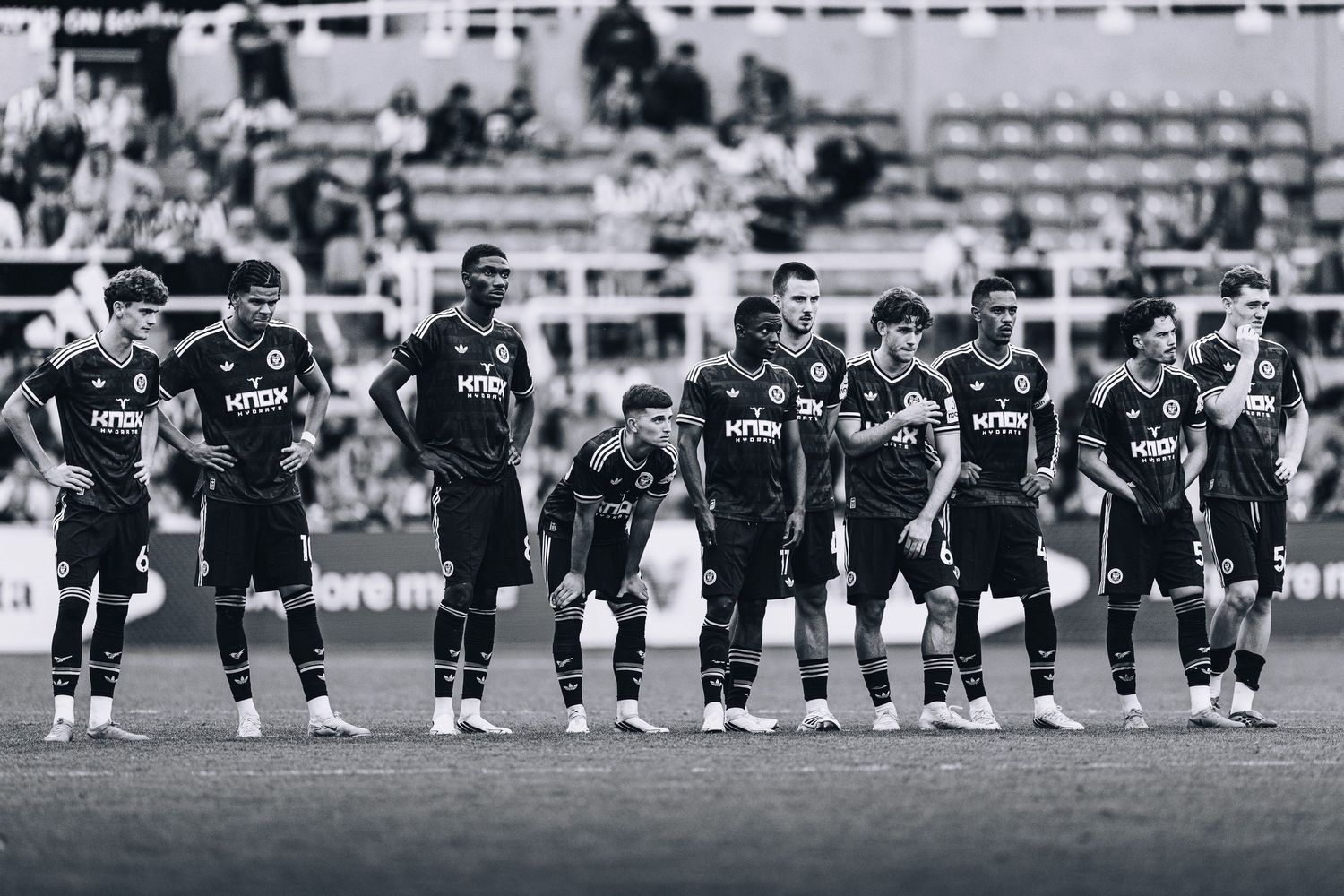 Black and white team photo of footballers lined up on pitch during game.