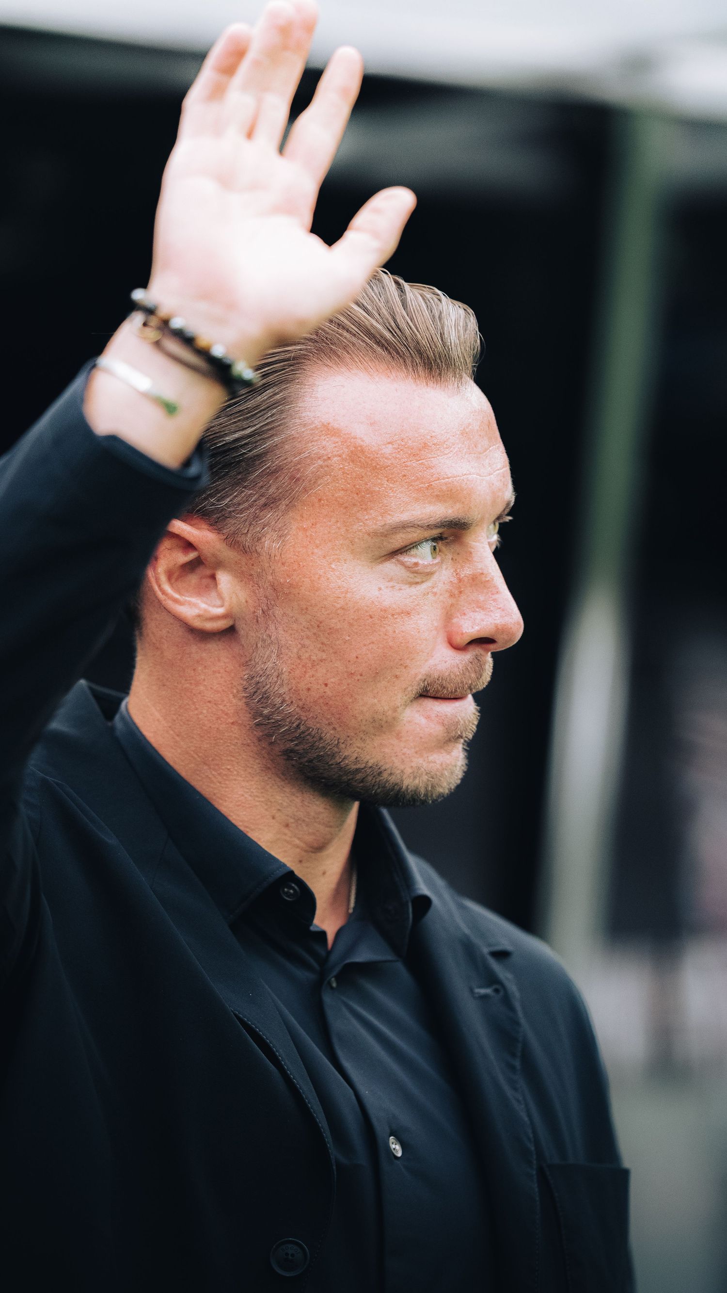 Blonde male coach in black shirt raises hand while looking sideways outdoors.