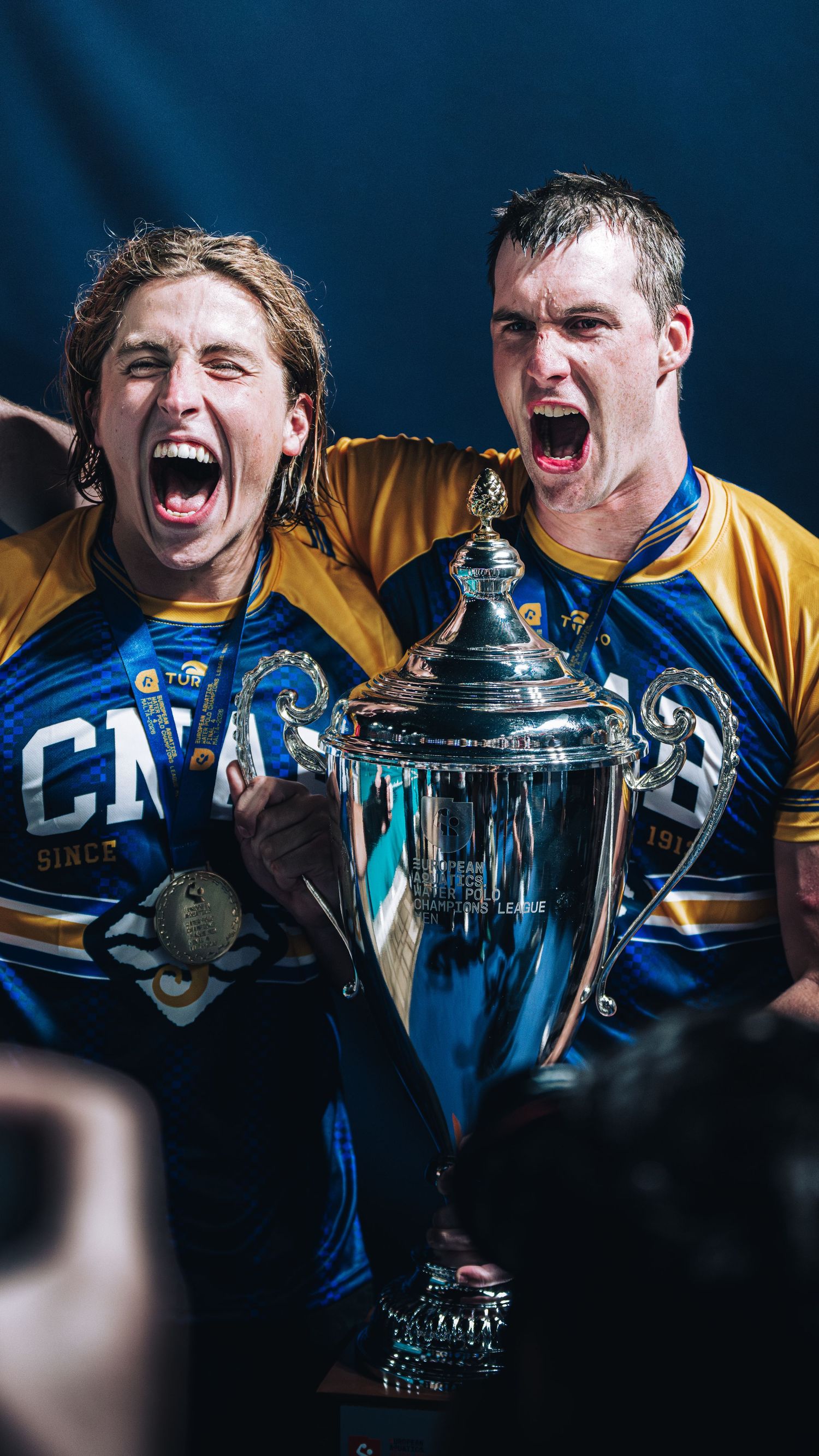 Two CNAB water polo players celebrating loudly while holding the champions trophy.