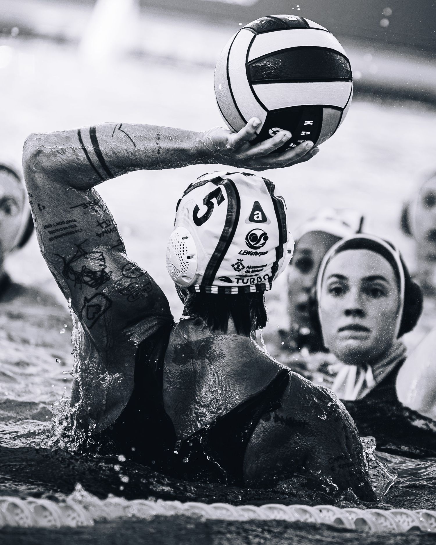 Black and white action shot of water polo player number 5 raising ball to throw during match.