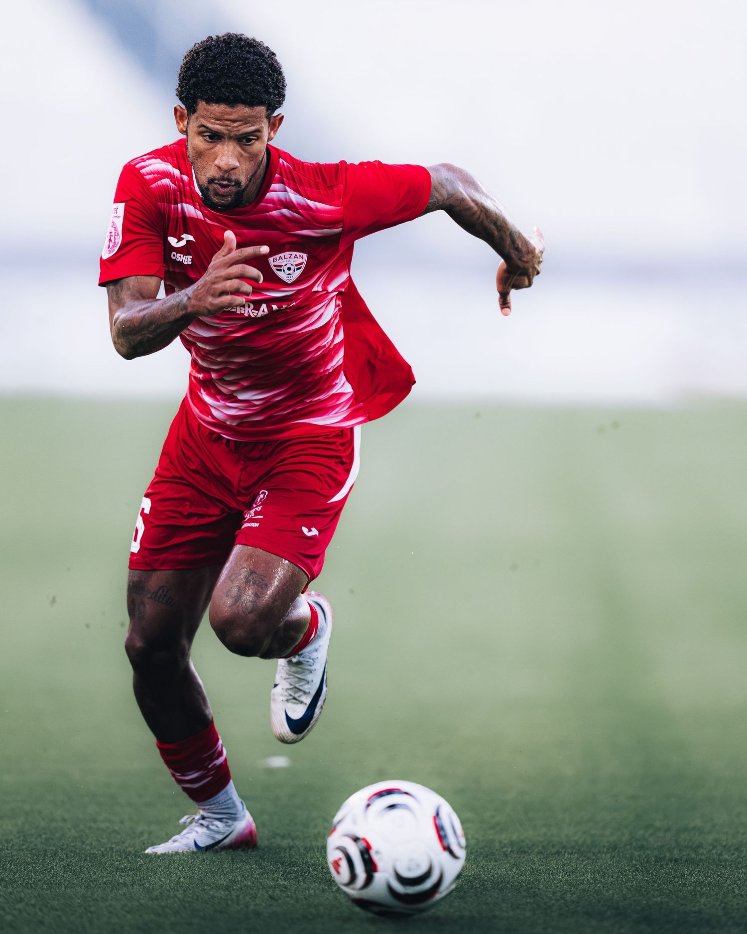Soccer player in red Joma kit sprints with ball during match.
