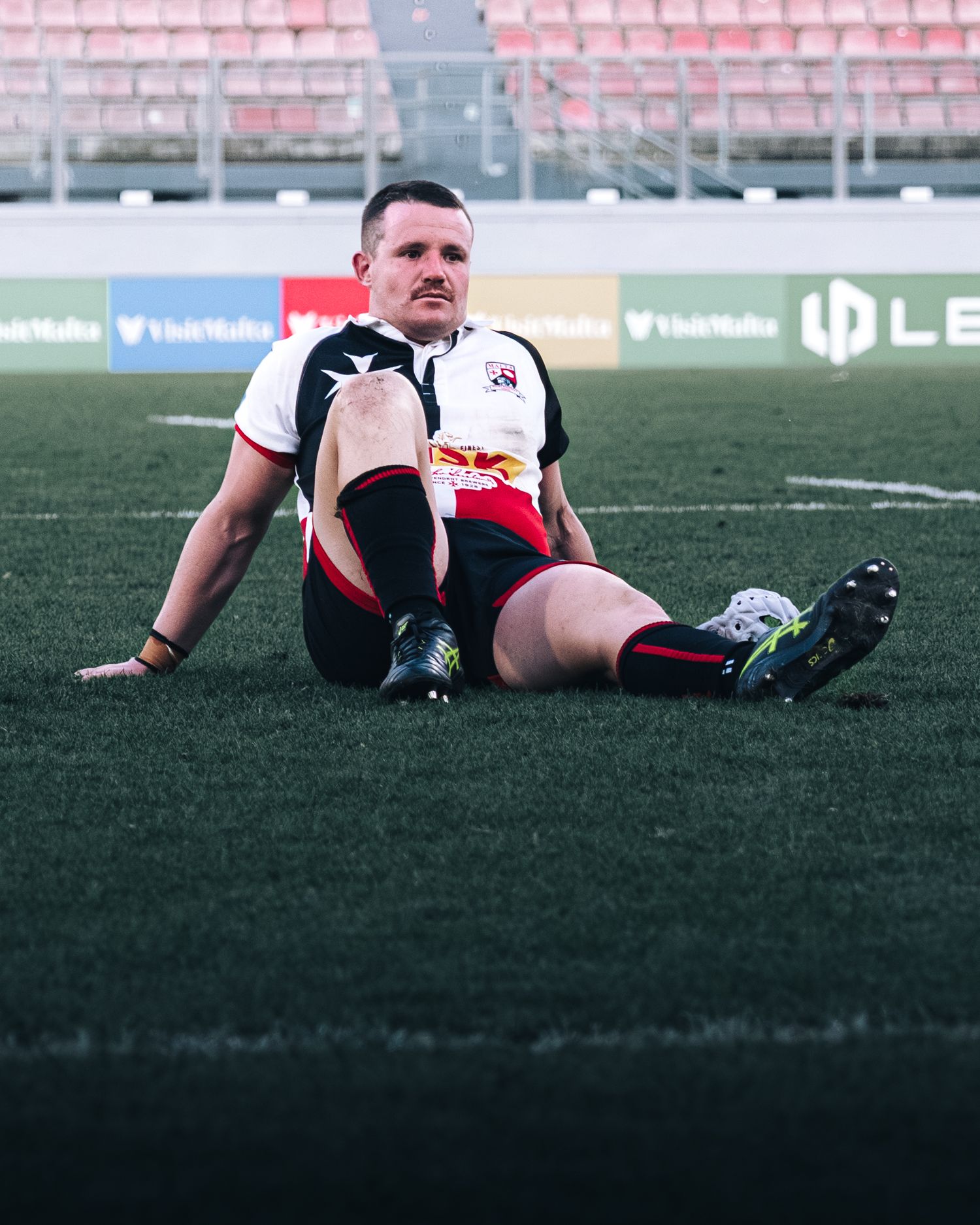 Rugby player sitting on grass field wearing black and white kit with red bull logo during match.