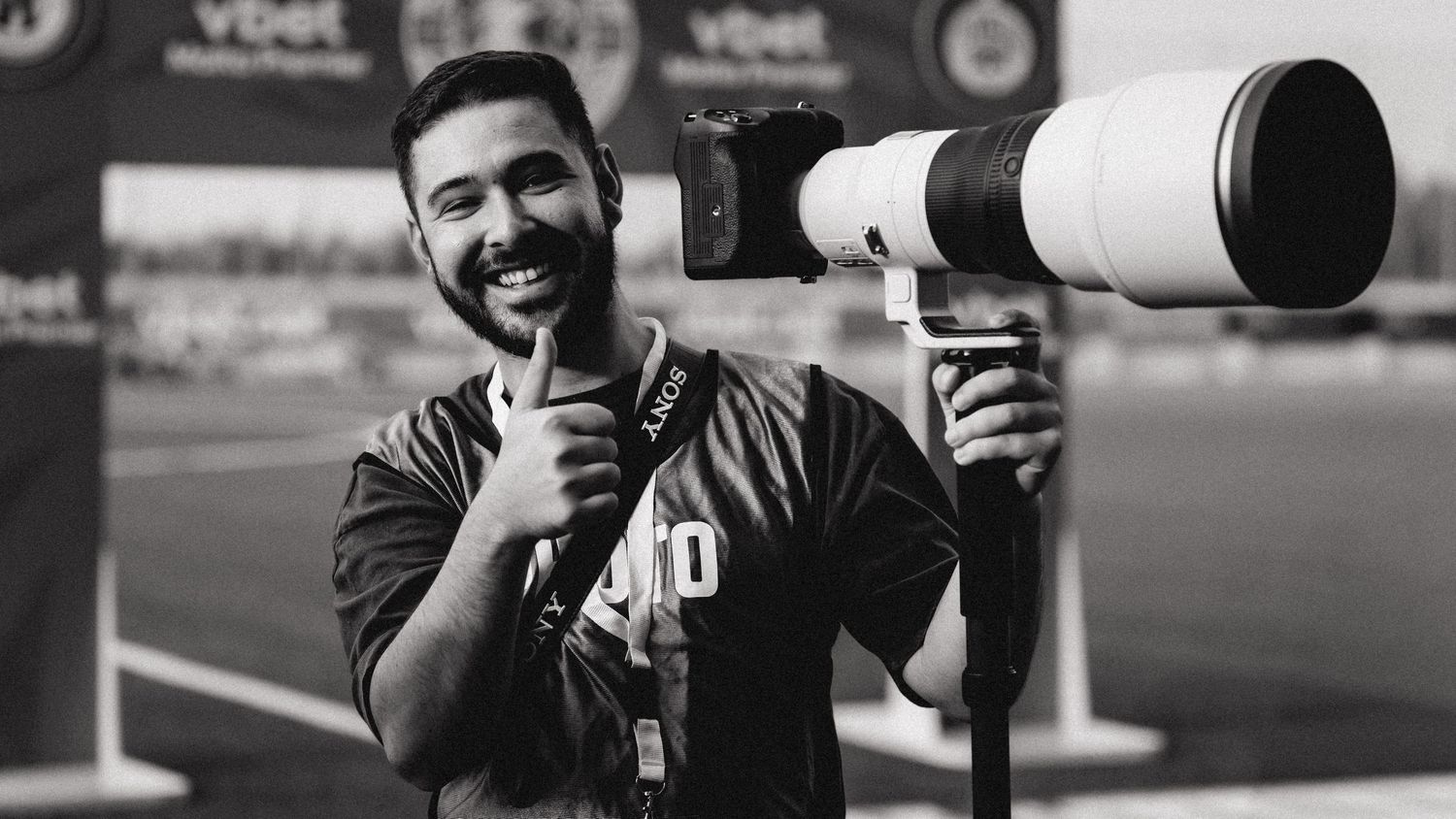 Black and white photo of a smiling sports photographer holding a large telephoto camera.