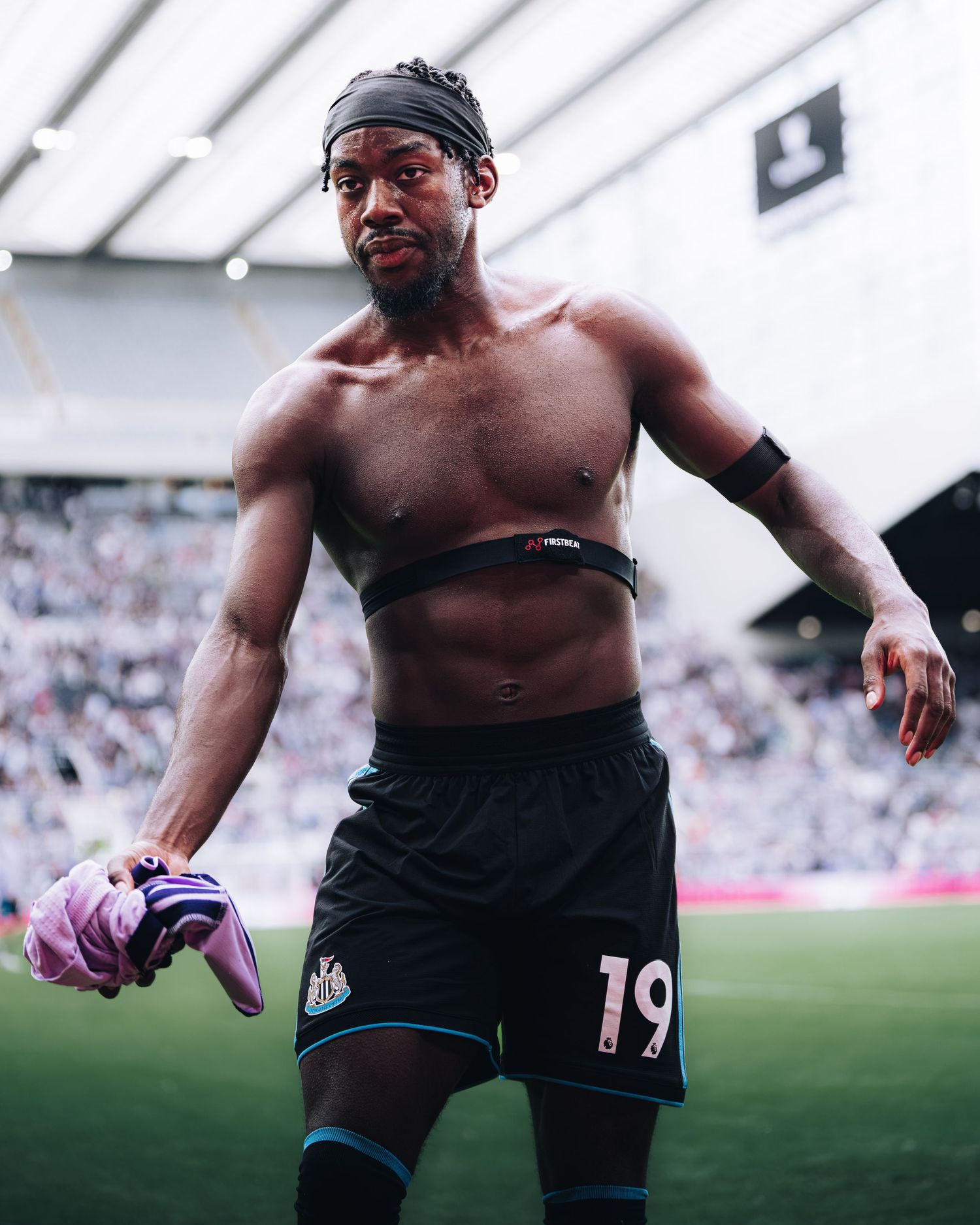 Shirtless soccer player wearing number 19 Newcastle shorts holding gloves on stadium pitch.