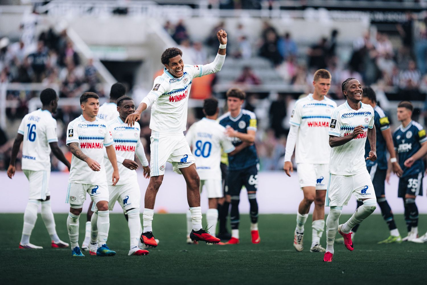White-uniformed soccer team celebrates on field while opponents in dark jerseys stand nearby.