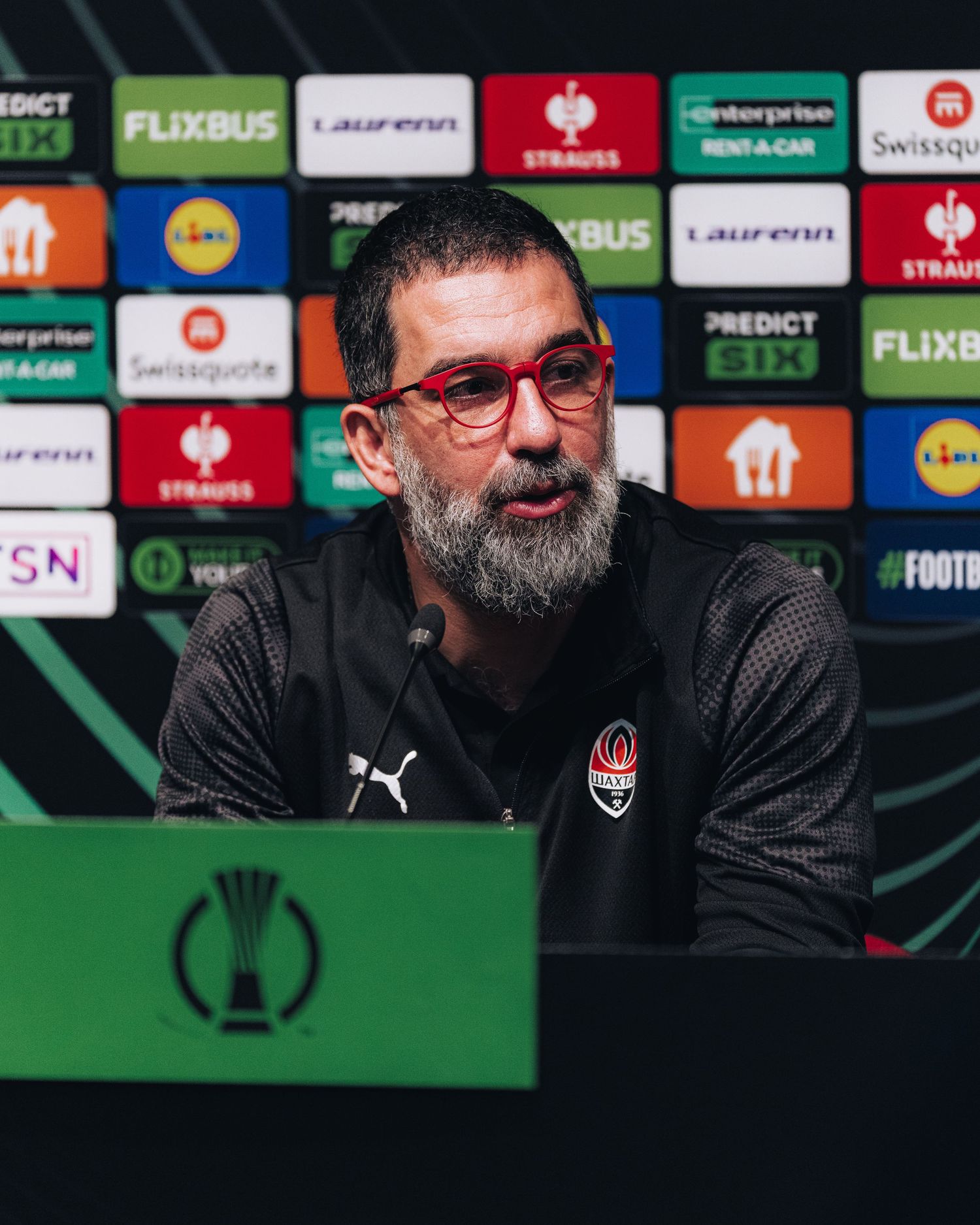 Bearded coach wearing red glasses speaking at UEFA Conference League press conference.