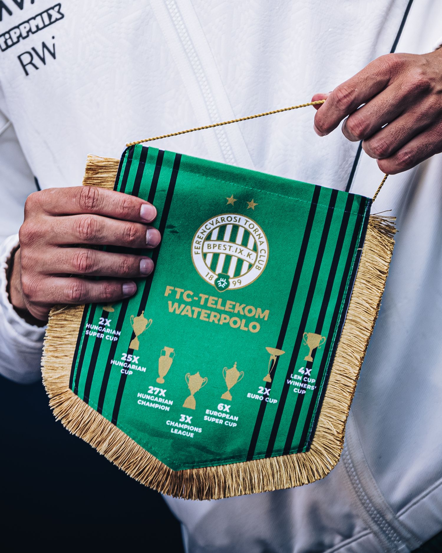 Hands holding green FTC-Telekom Waterpolo pennant with gold fringe showing club achievements and trophies.