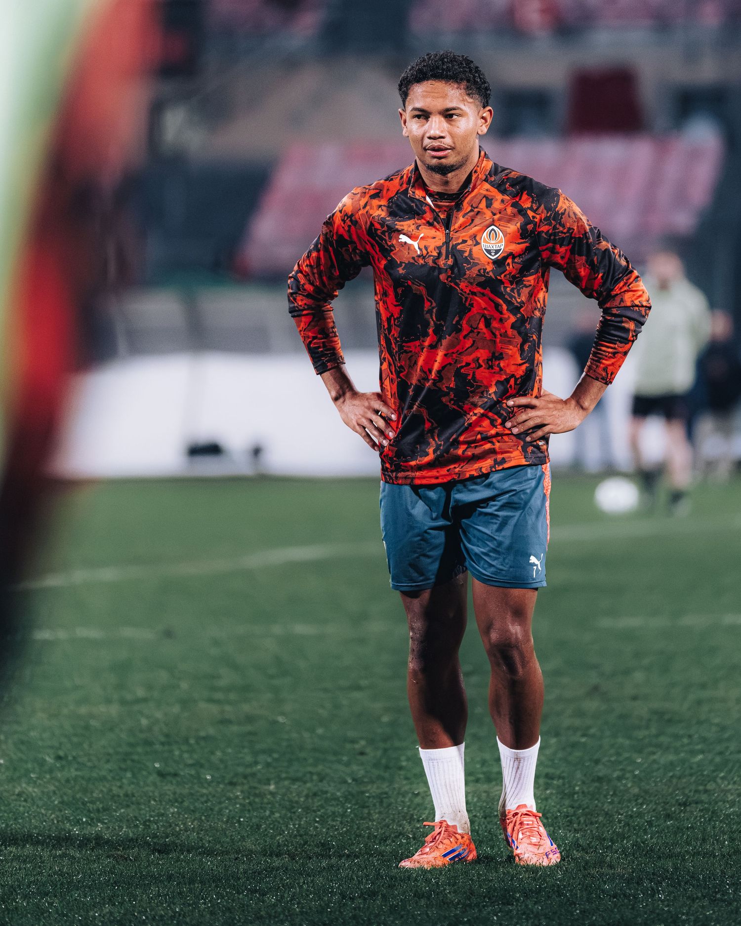 Soccer player in orange PUMA jersey with hands on hips standing on green field during warm-up.