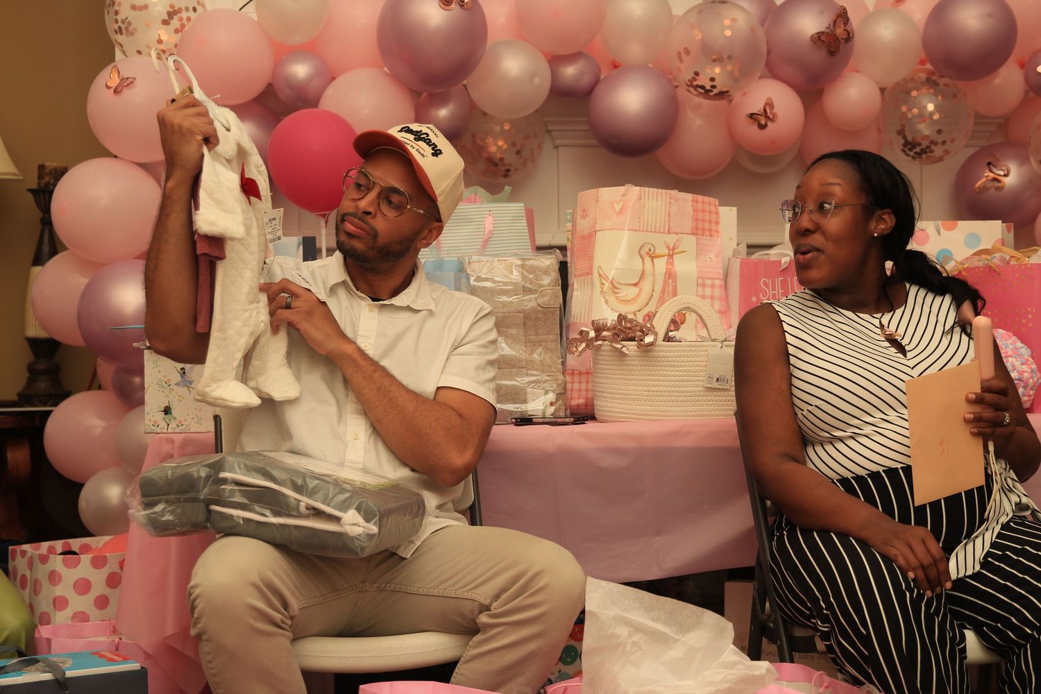 Man holding up white baby socks at a pink-themed baby shower with balloon decorations.