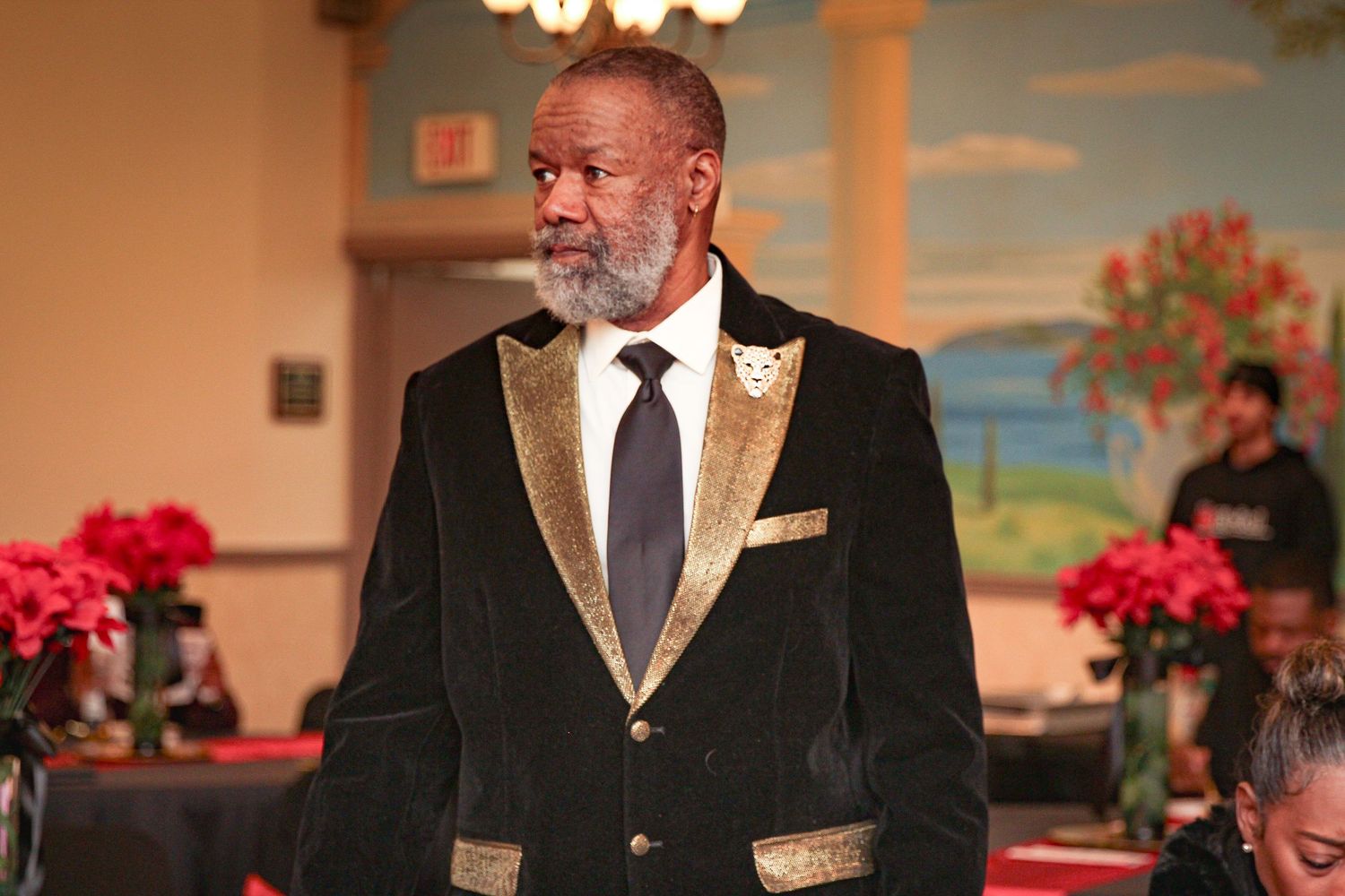 Elegant man in black velvet tuxedo with gold trim and blue tie at formal event with red floral decor.