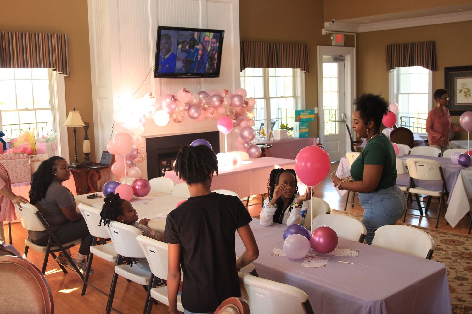 Children and adults gather at a pink balloon-decorated birthday party in a warmly lit banquet room.