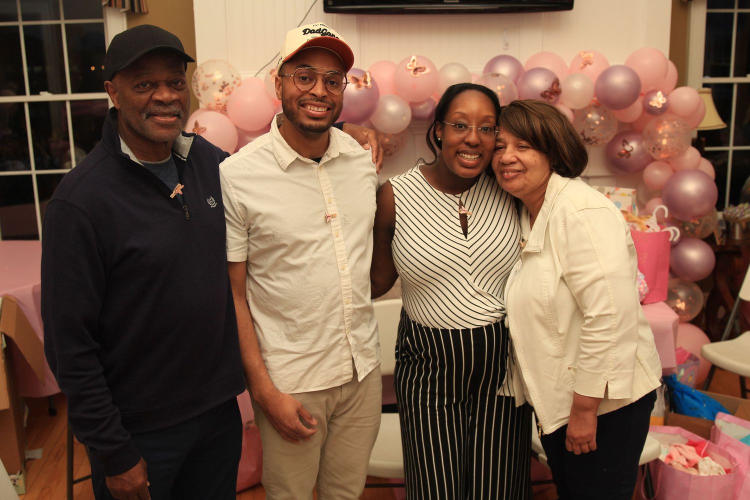 Four people smile together at a pink baby shower celebration with decorative balloon arch background.