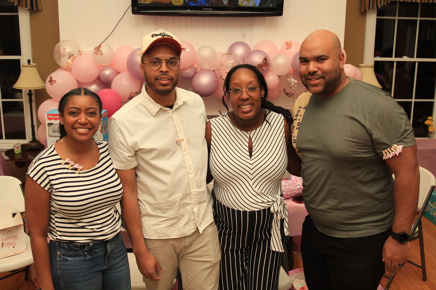 Four people pose together smiling at a pink-themed baby shower with balloon arch decoration.