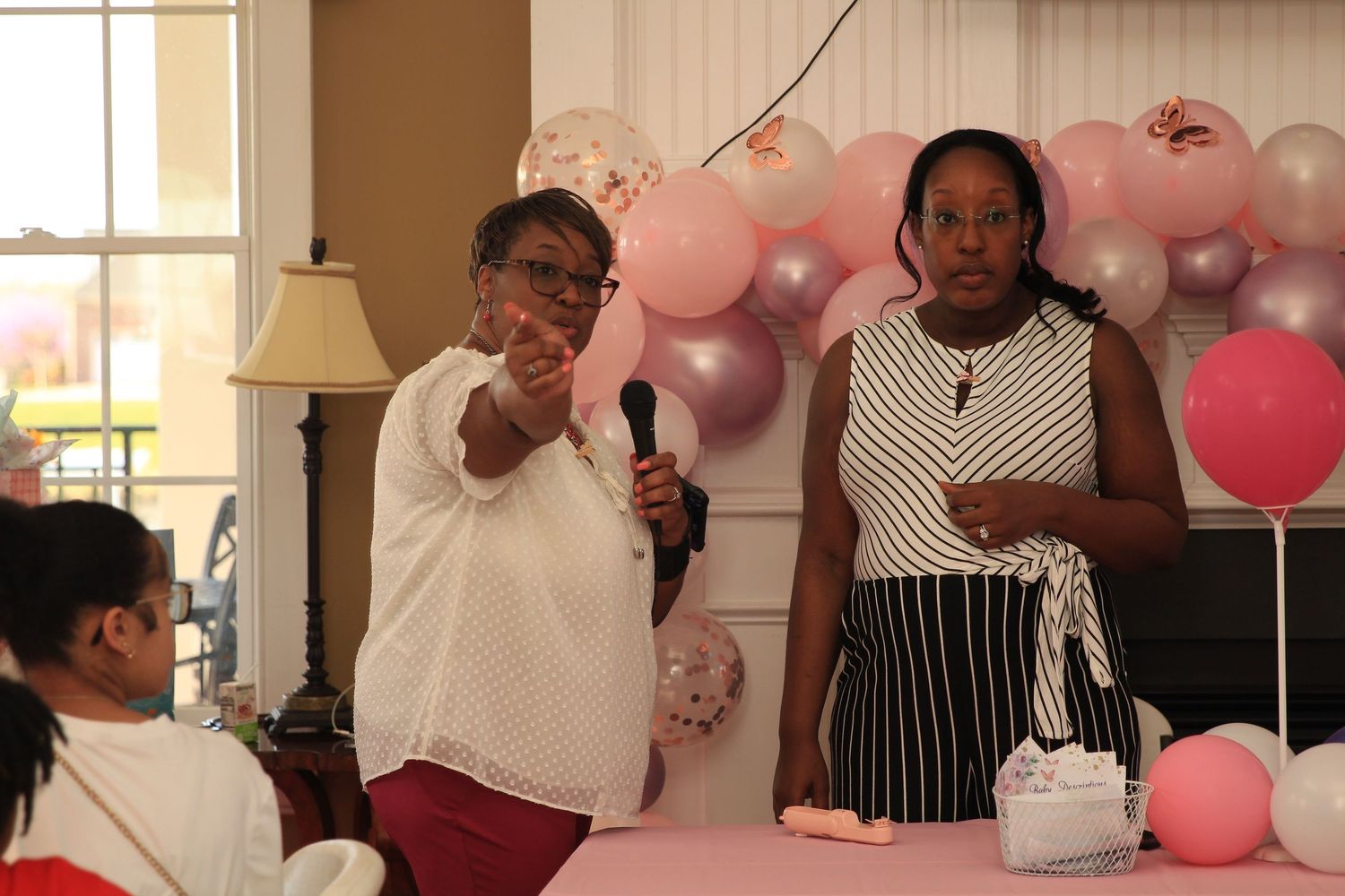 Two women speak with a microphone at a pink baby shower event.