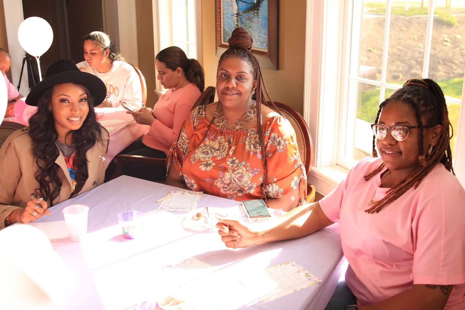Three women smile at a table during a pink-themed baby shower celebration.