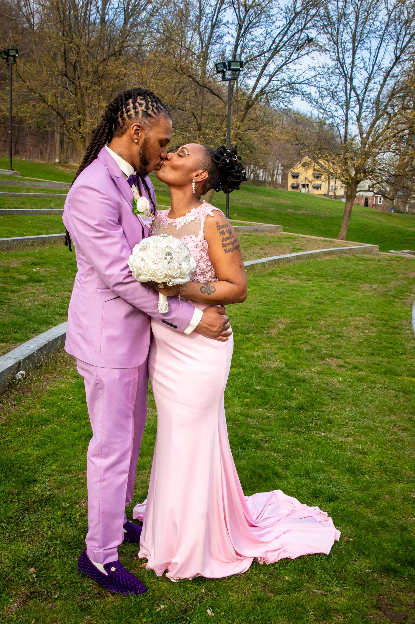Couple kissing outdoors in matching pink wedding attire, groom in lavender suit and bride in pink gown.