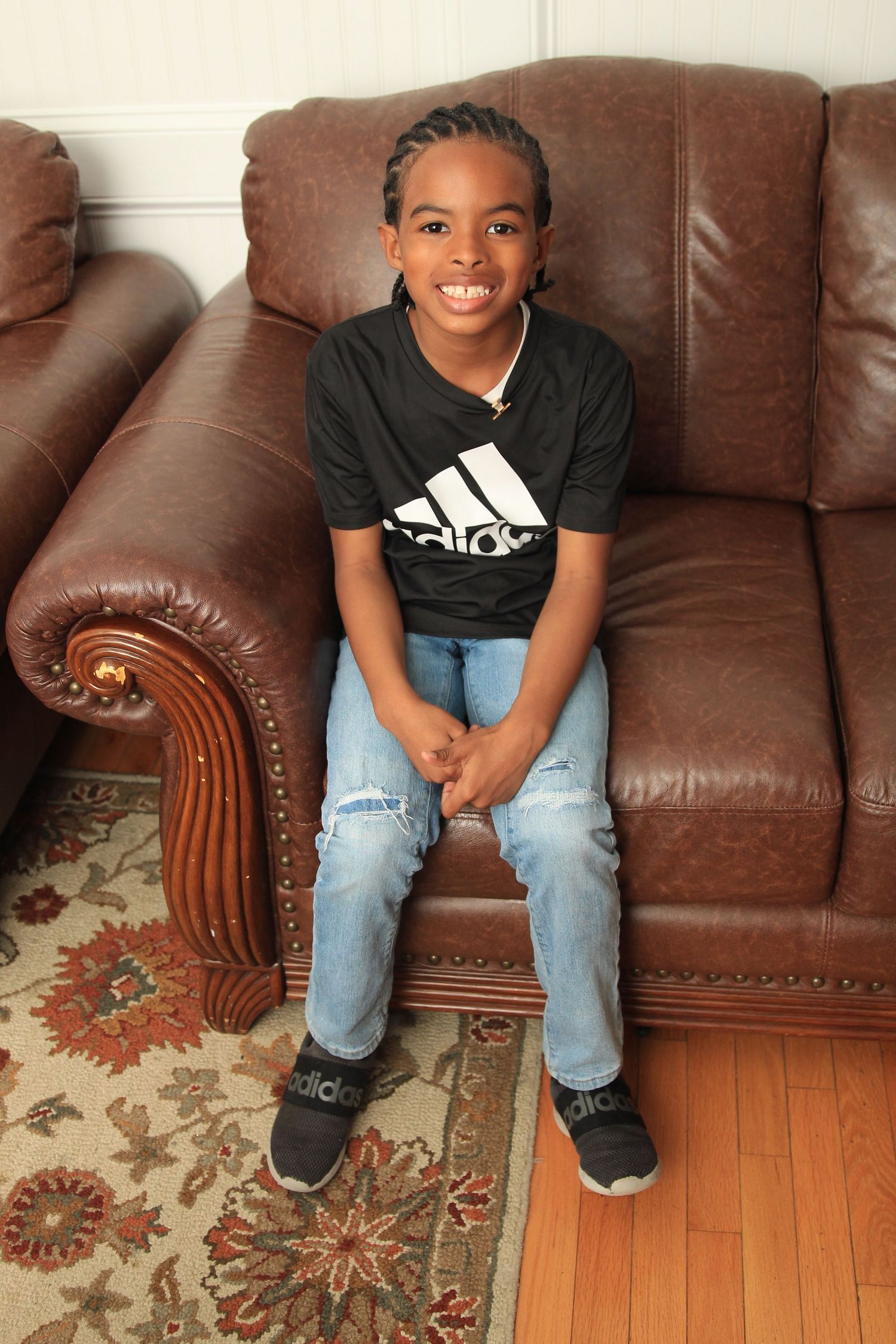 Smiling young boy wearing an Adidas shirt sitting on a brown leather couch.