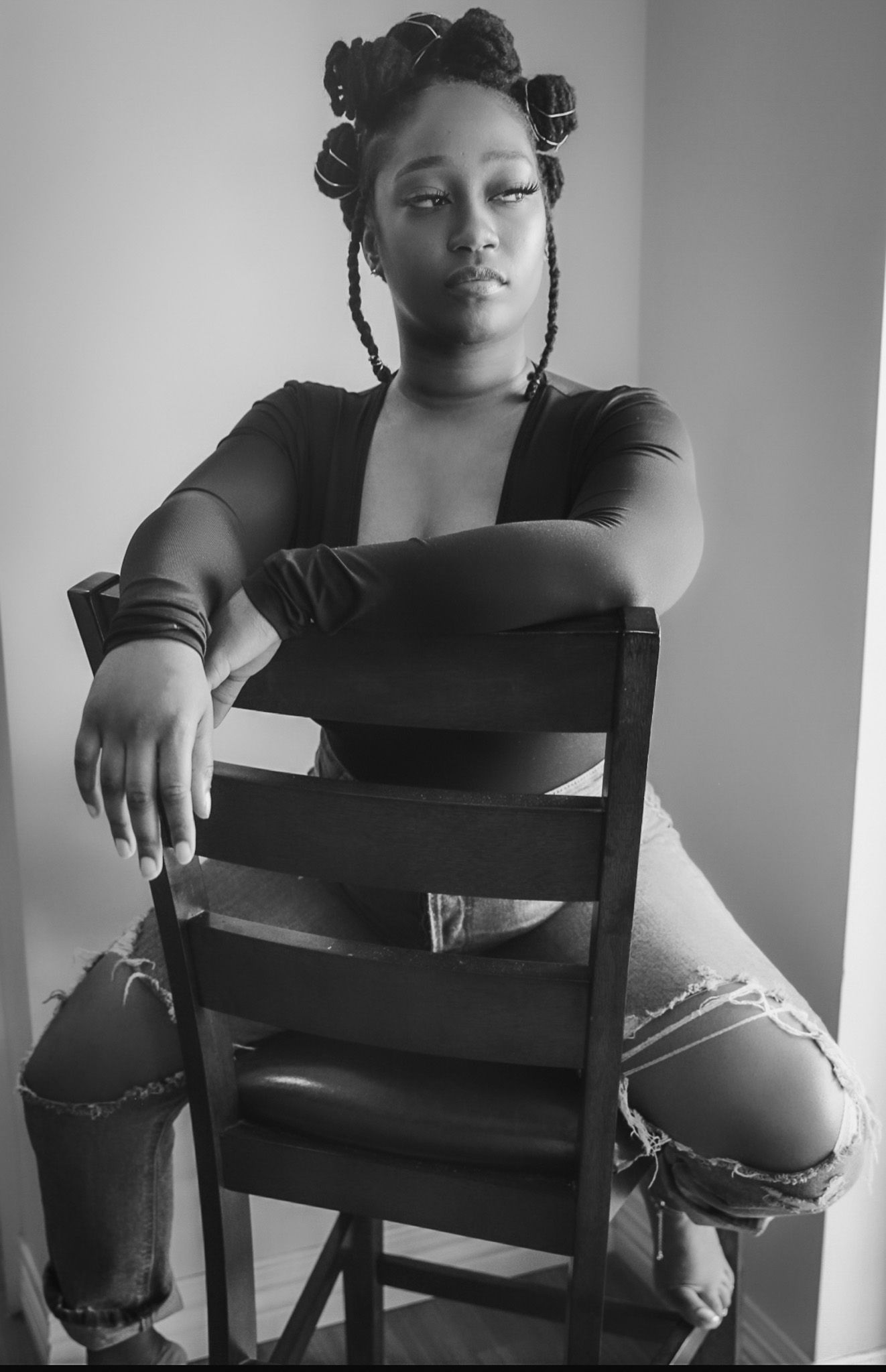 A young woman poses on a wooden chair in black and white photo, wearing ripped jeans and black top.
